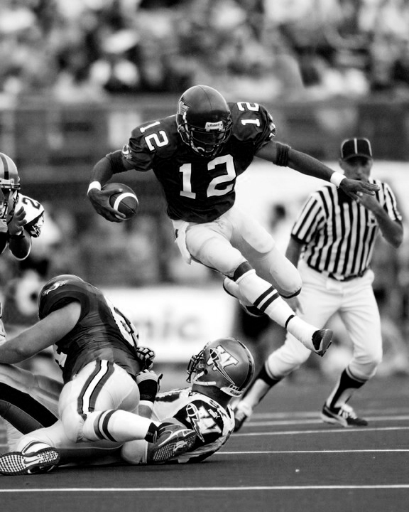 Quarterback on the field running with the ball wearing a number 12 jersey. A referree is in the background as well as a few players on the ground.