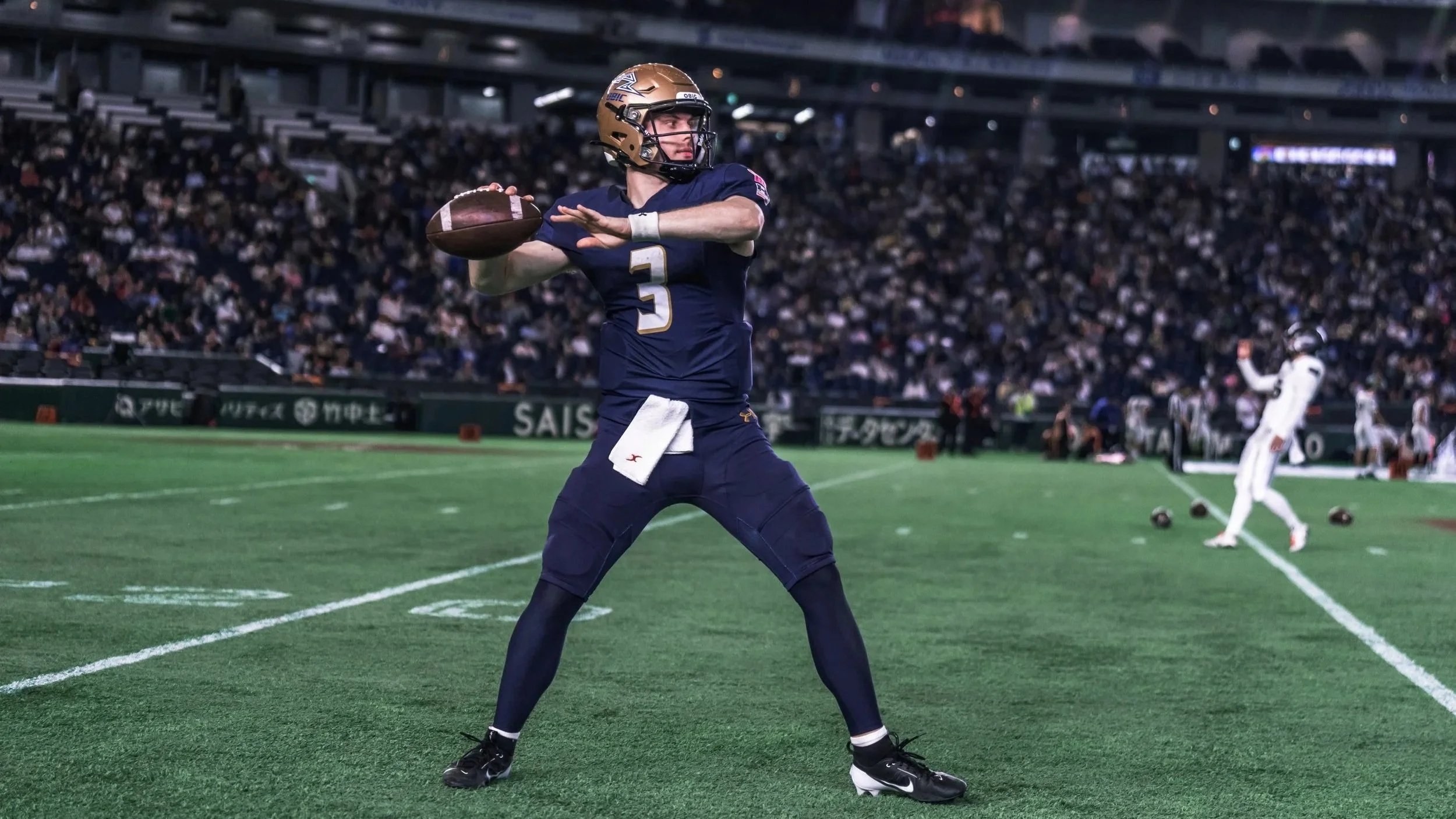 American football player in navy uniform with gold helmet and white towel, preparing to throw the ball on a football field at night, with a crowd in the stands and other players in the background.