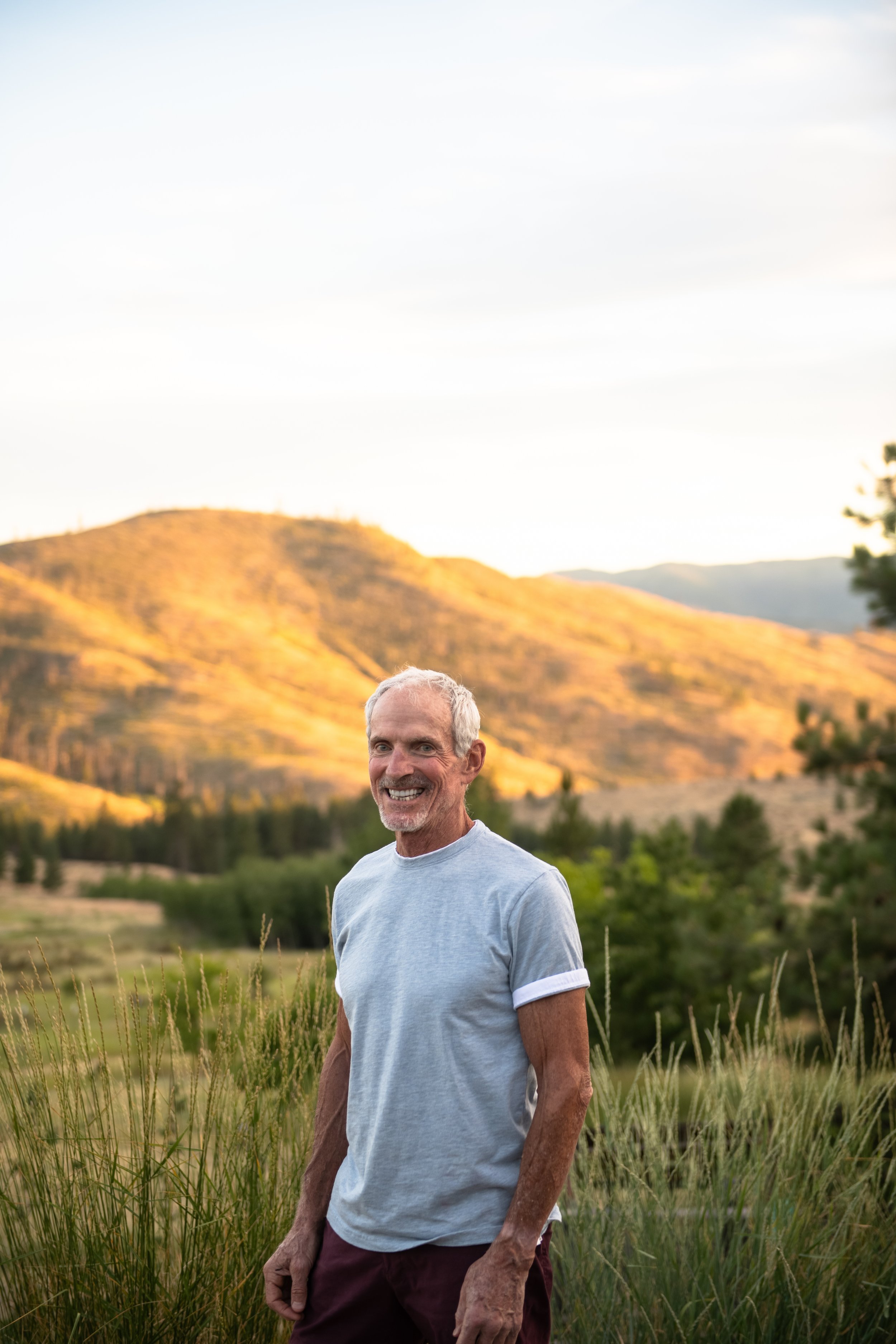 A smiling Rob Spence standing outdoors in a grassy field, with hills and trees in the background during sunset.