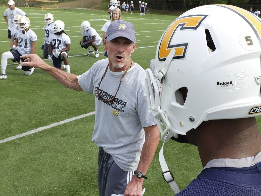 Rob Spence gives instructions to a University of Tennessee Chattanooga Mocs player wearing a white helmet with a yellow 'C' logo during a practice on a football field, with other college players in the background.