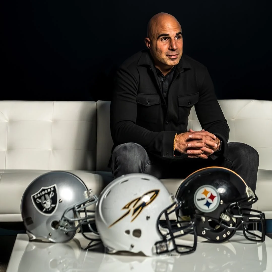 Rob Gronkowski sitting on a white couch with his hands clasped, three football helmets on a table in front of him, with a dark background.