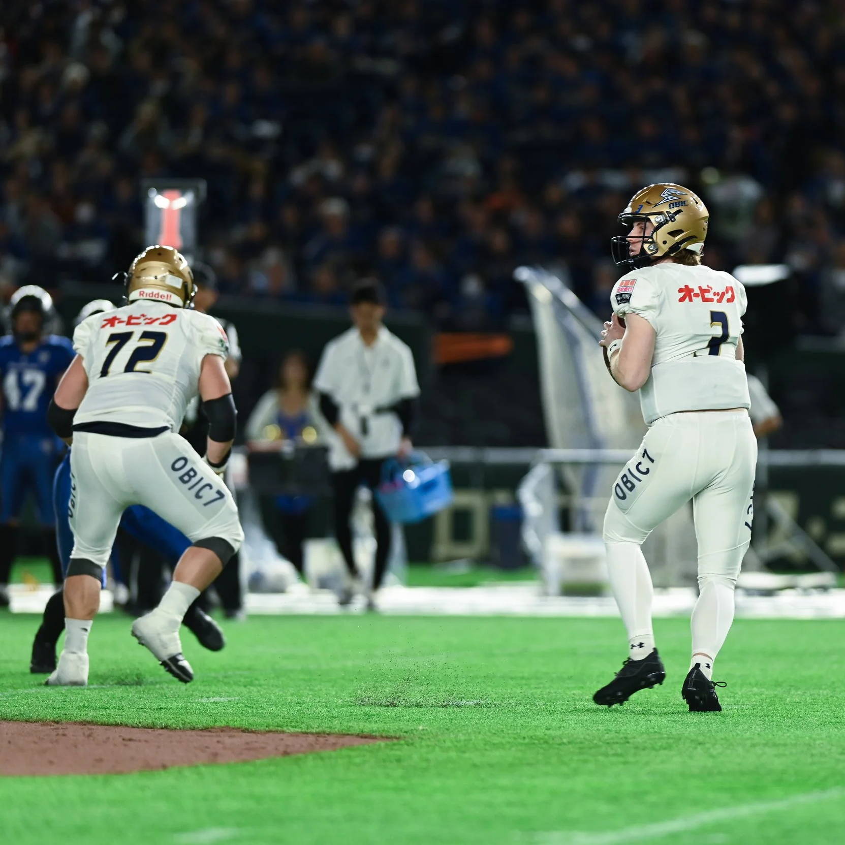 American football players in white uniforms with Japanese characters on a green field, with a crowd in the stands.