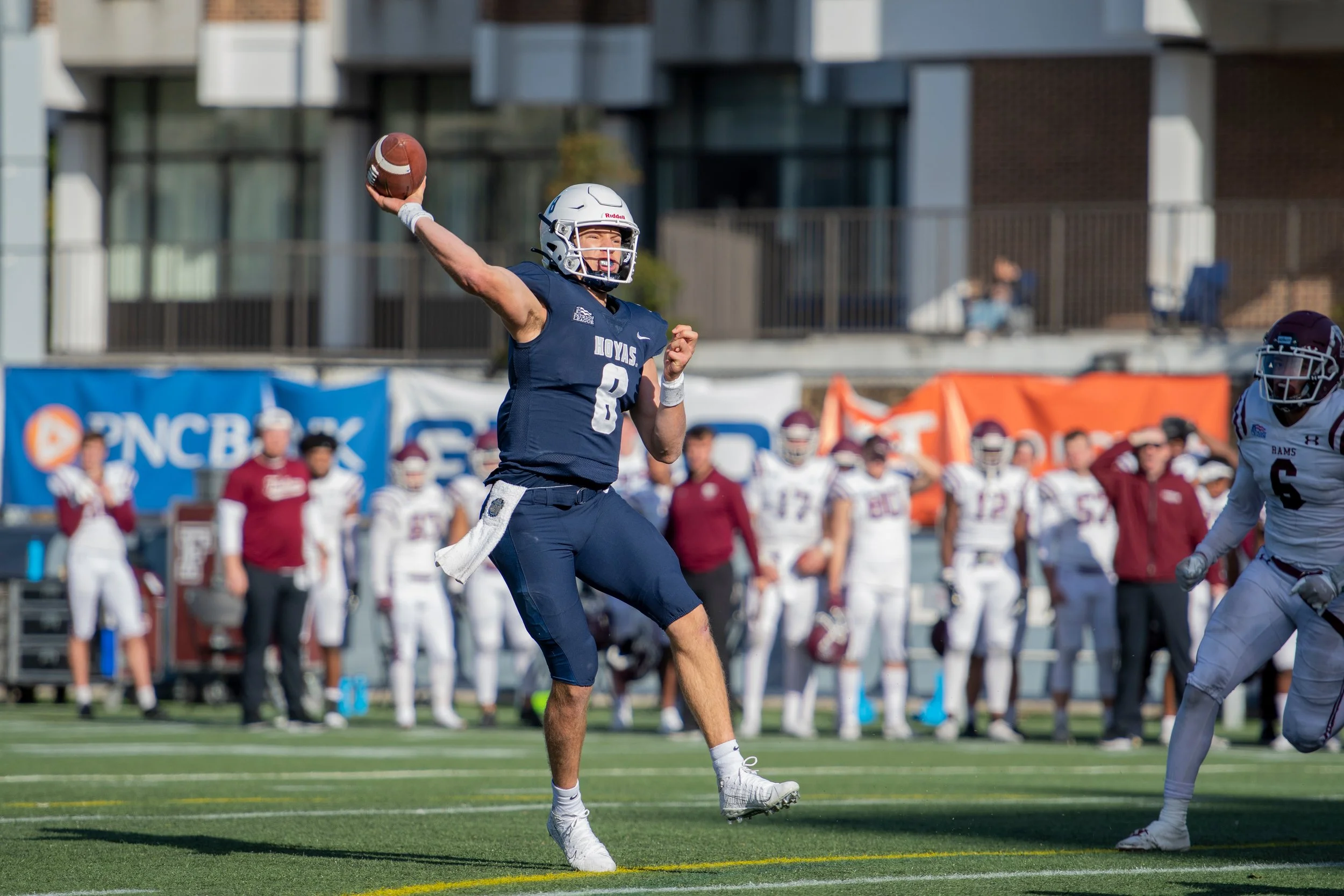A quarterback mid throw wearing a blue Hoyas uniform with the number 8 and a white helmet and shoes with players on the sideline in the background.