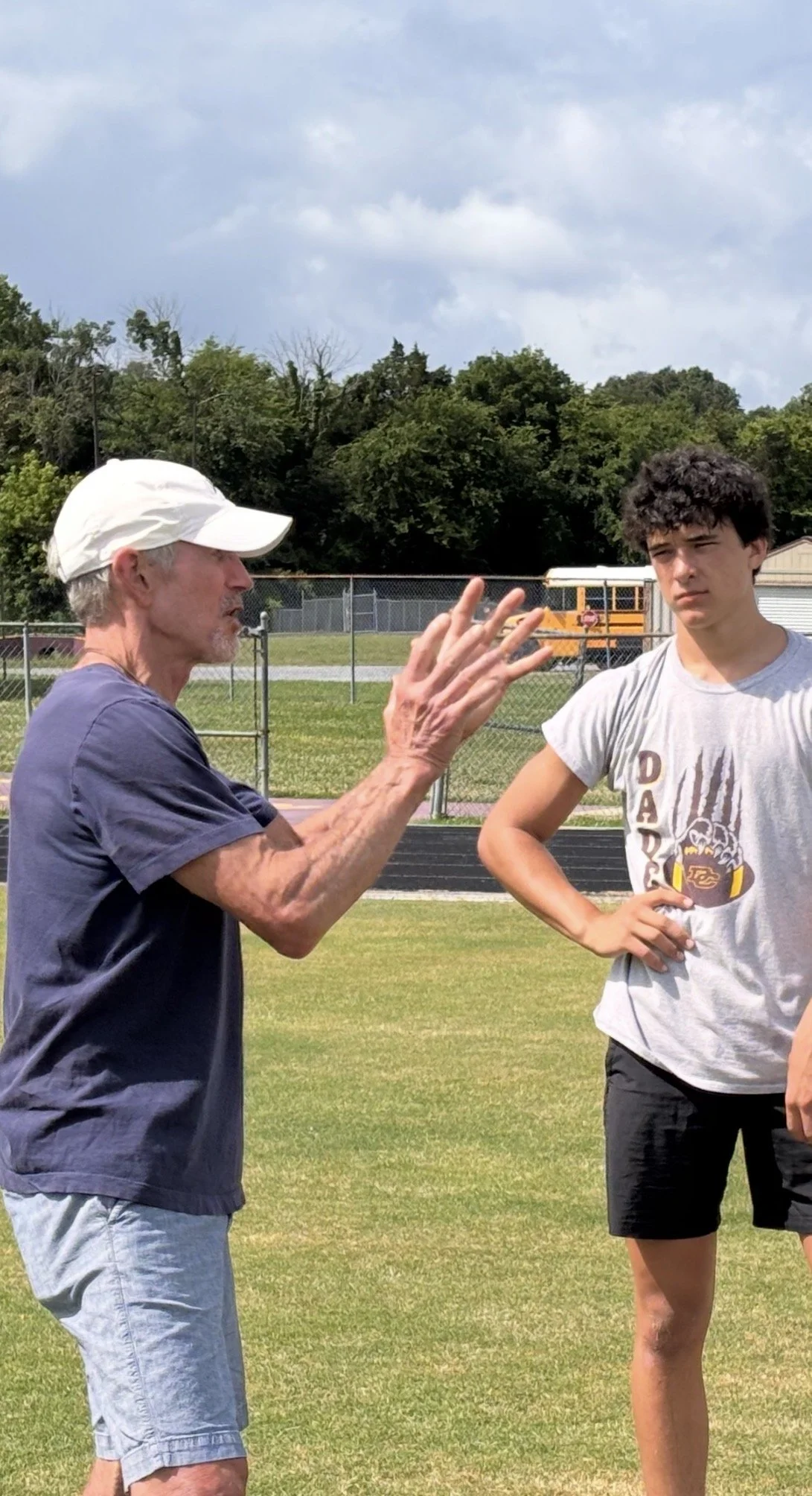 Coach Spence speaking to a teenage boy with curly dark hair at a school football field. Coach is gesturing with his hands, and the boy has his hand on his hip, listening.