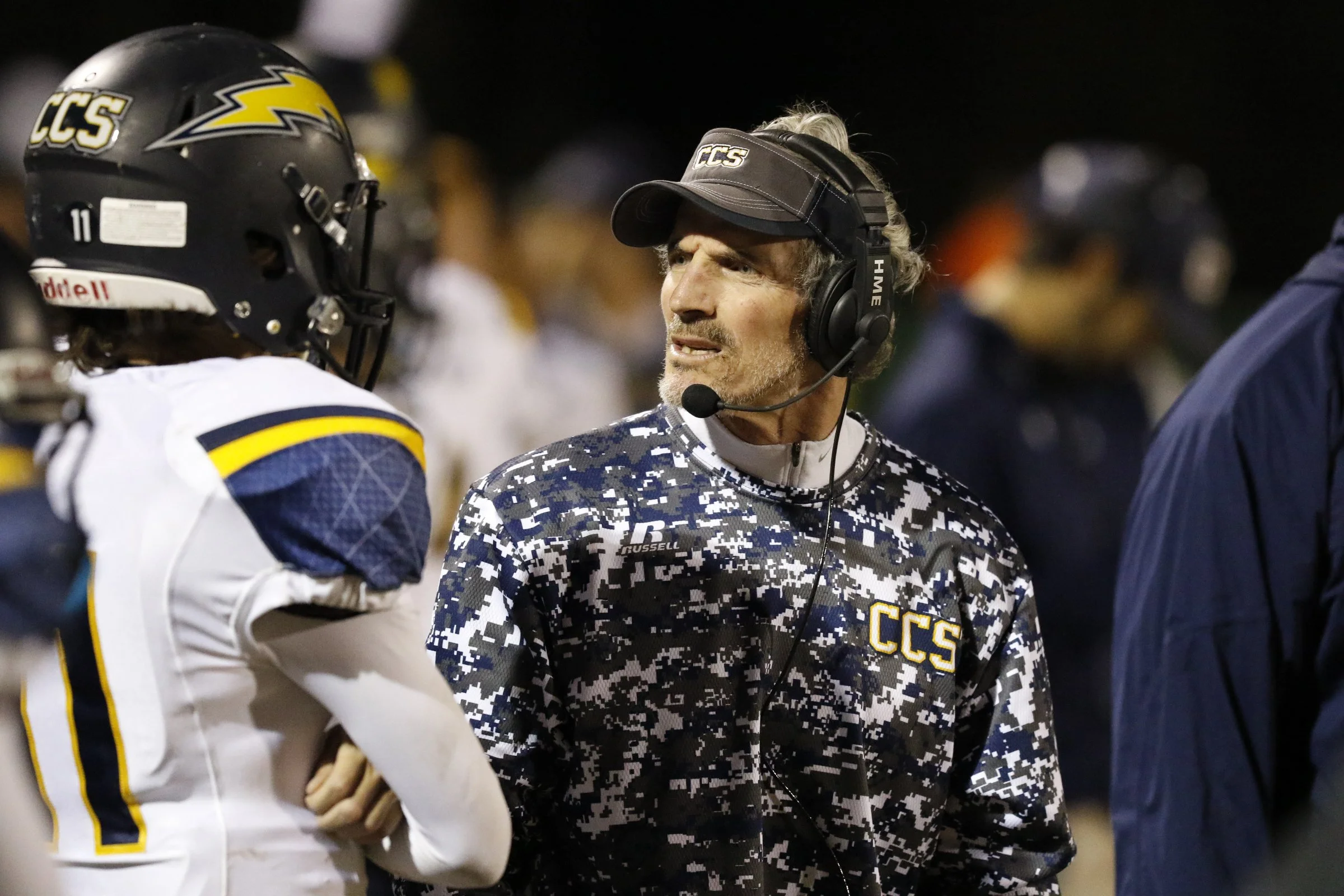 A football coach wearing a camouflage-patterned jacket and headset speaking to a player, both on the sidelines of a game at night. The player is dressed in a football uniform with a helmet.