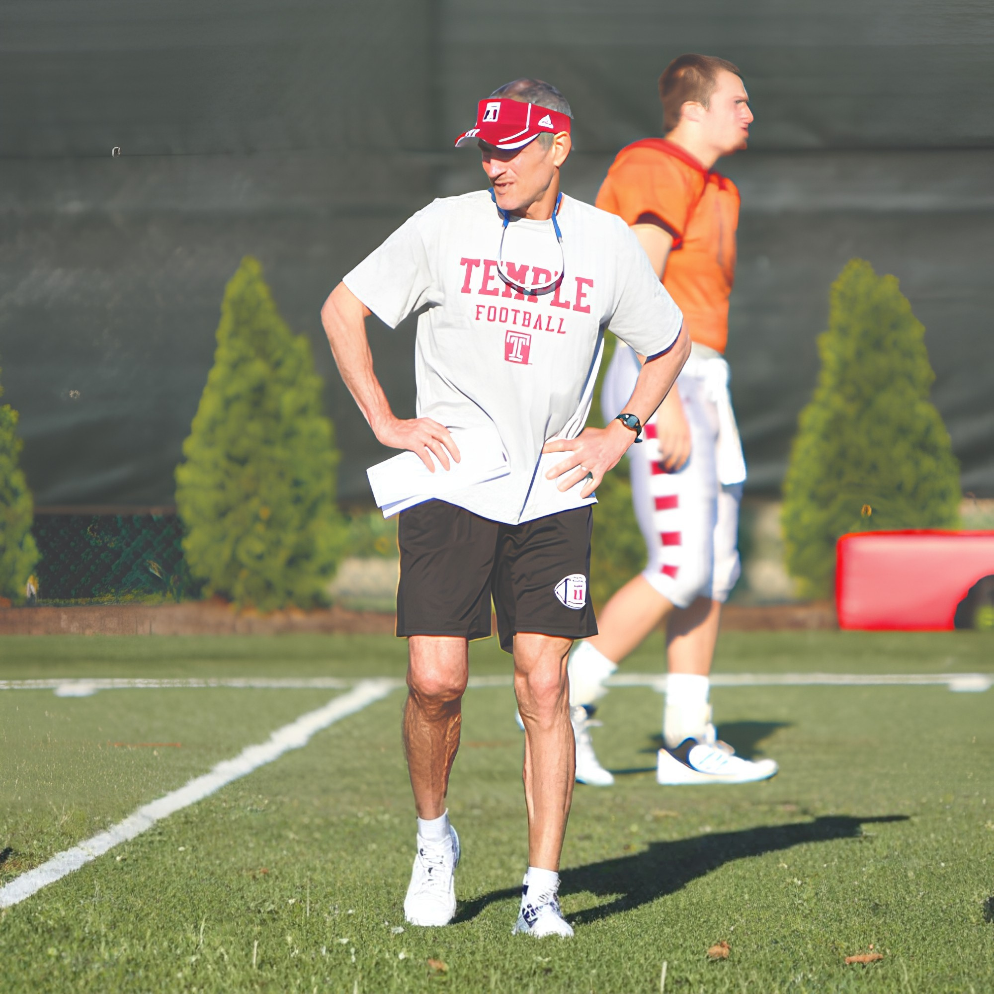 Coach Spence in a white T-shirt with red text that says Temple, black shorts, white sneakers, and a red hat stands on a grassy field during daylight, with another man in an orange shirt and white shorts visible in the background.
