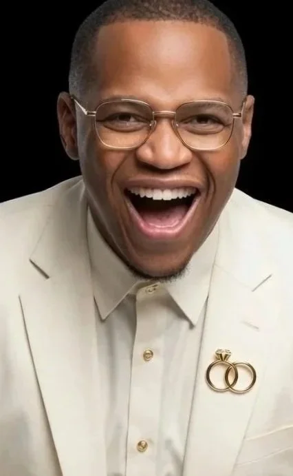 Man smiling and laughing, wearing glasses and a light-colored suit with wedding rings pines on his lapel.