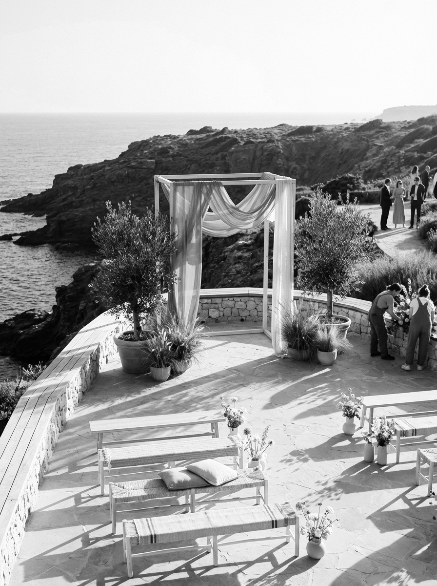 A black-and-white photograph of an outdoor wedding ceremony setup on a terrace overlooking the ocean, with an arch draped in fabric, benches, flower arrangements, and a small group of people near a stone wall.