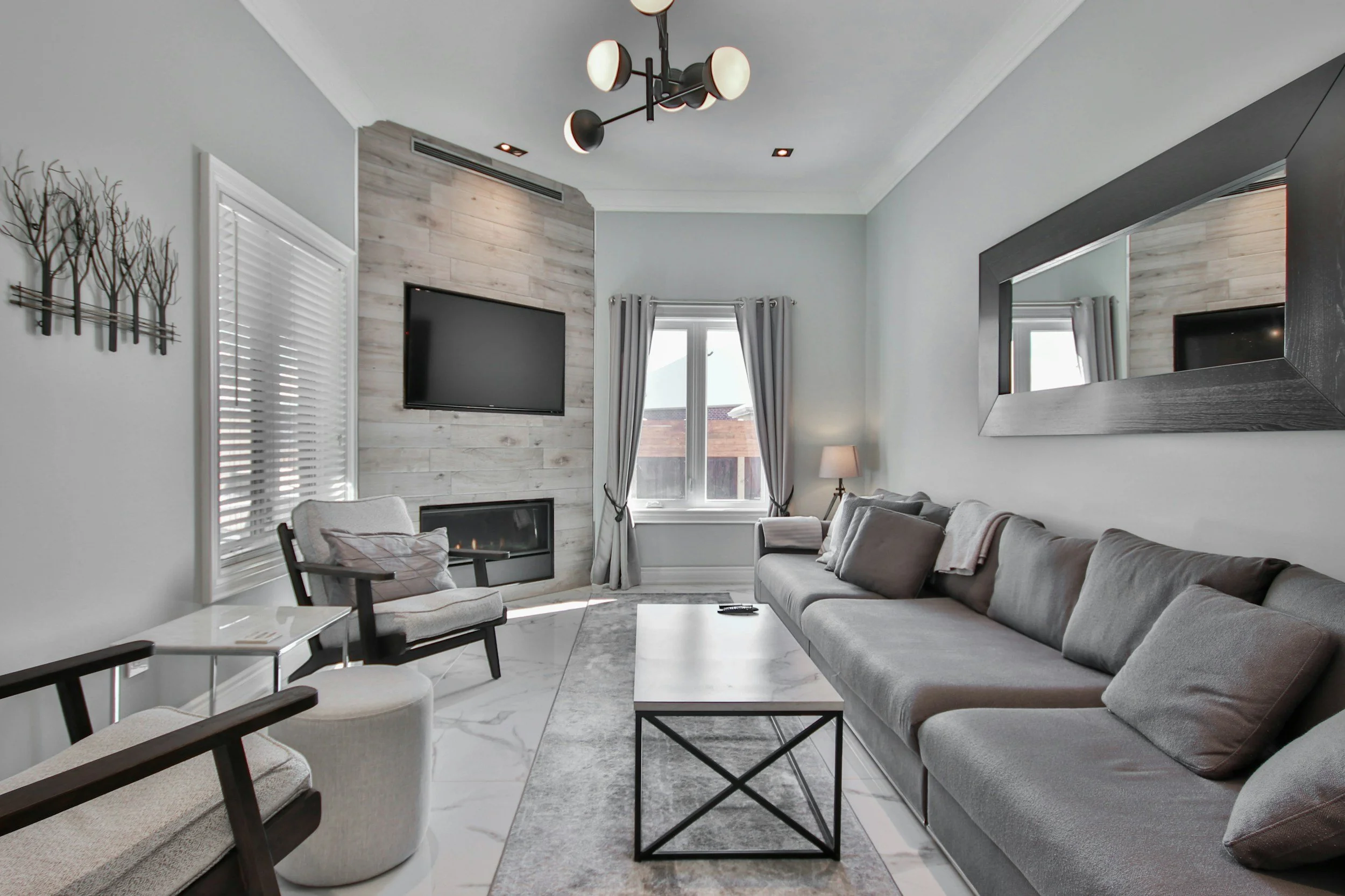 Modern living room with a gray sectional sofa, a white marble coffee table, black armchair with beige cushions, wall-mounted TV, large window with curtains, framed mirror, and a ceiling light fixture.