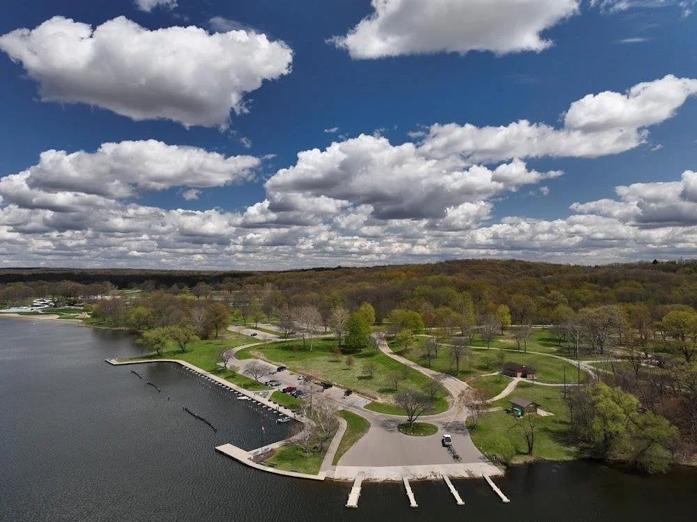 Aerial view of a lakeside park with walking trails, trees, and a small dock with boat slips under a cloudy sky.