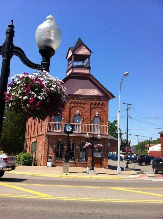 A historic brick building with a clock and a small tower on top, located on a street corner with hanging flower baskets and streetlights.