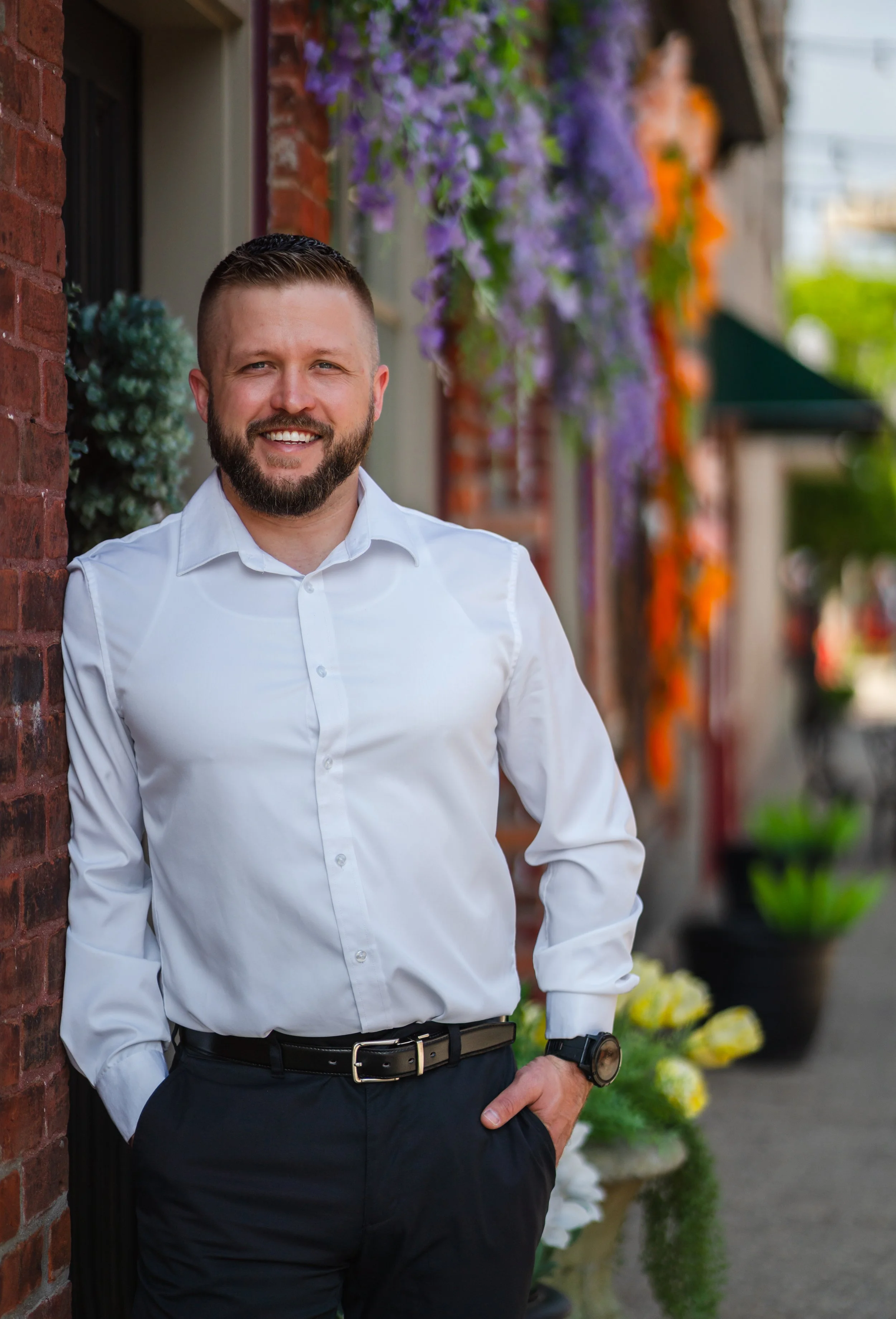 A man dressed in a white button-up shirt and black pants, with a beard and short hair, standing outdoors next to a brick wall with purple and orange flowers in the background.