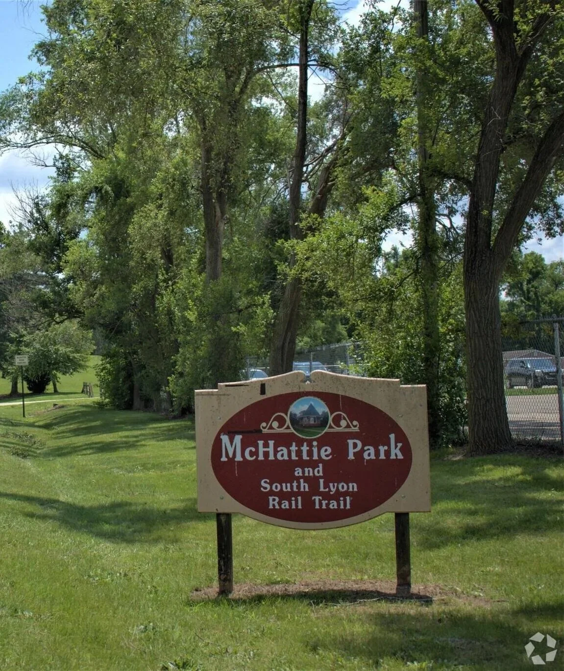 Sign for McHattie Park and South Lyon Rail Trail in a grassy park area with trees and a fence in the background.