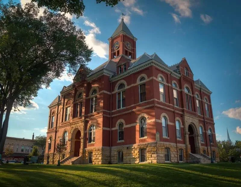 Historic red brick courthouse with clock tower, arched windows, and stone foundation, surrounded by green grass and trees under a partly cloudy sky.