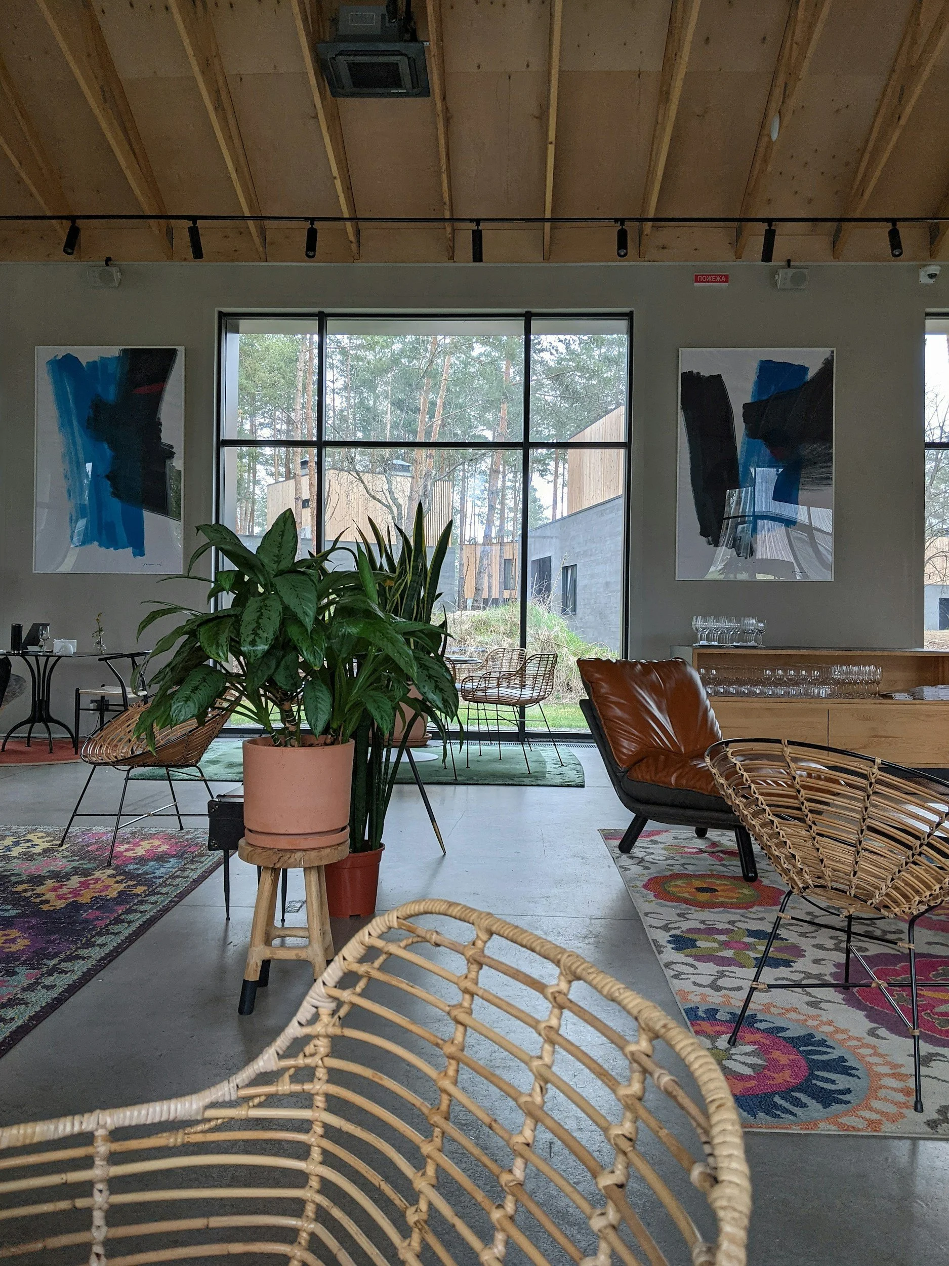 Interior of a modern room with a large window showing trees outside. The ceiling is made of exposed wood beams with track lighting. The room has two abstract paintings on the wall, a plant in a pink pot, and various chairs including wicker and leather ones. A patterned rug is on the floor.