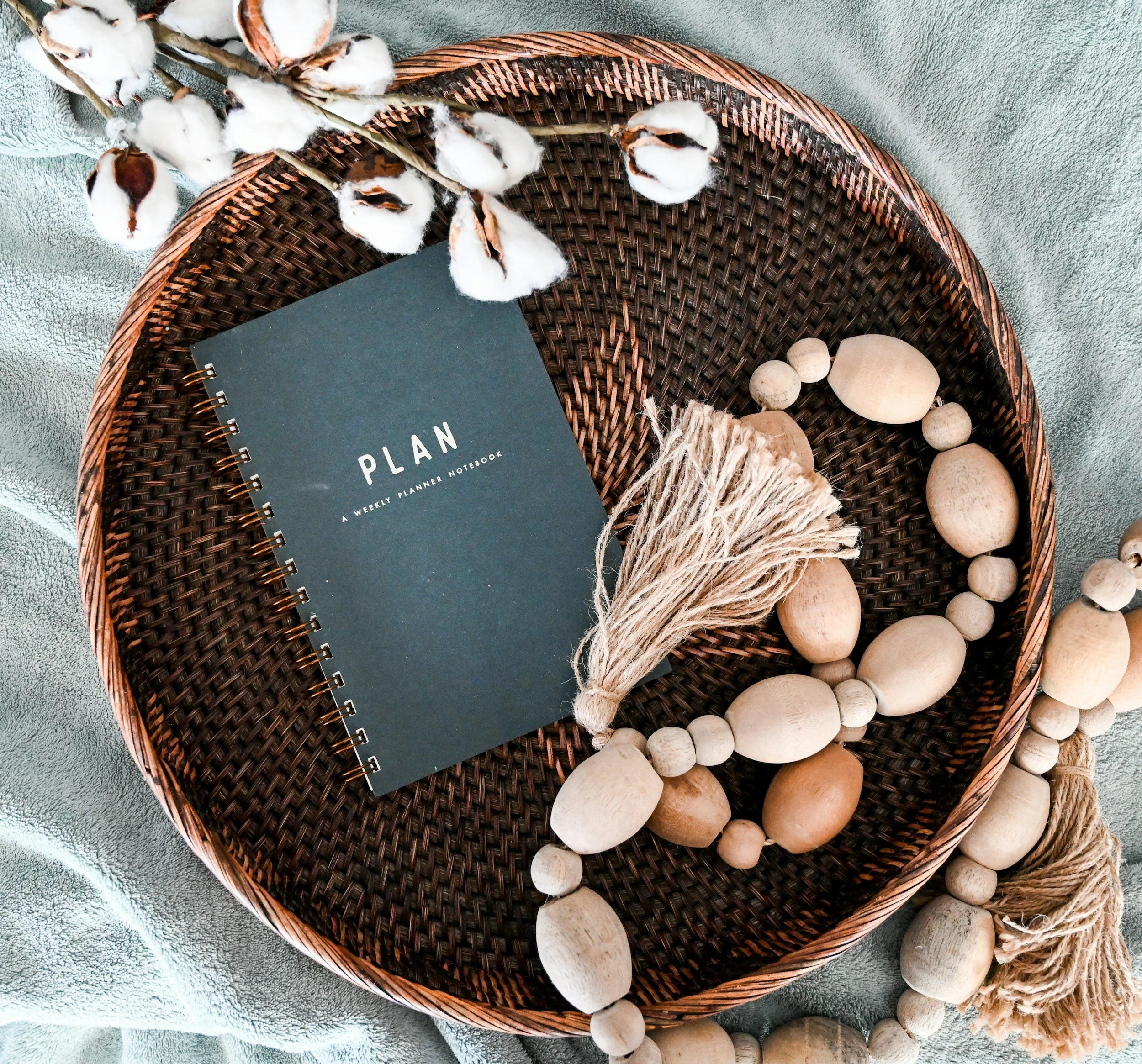 A woven basket containing cotton branches with cotton bolls, surrounded by wooden beads and a black planner notebook on a textured gray surface.