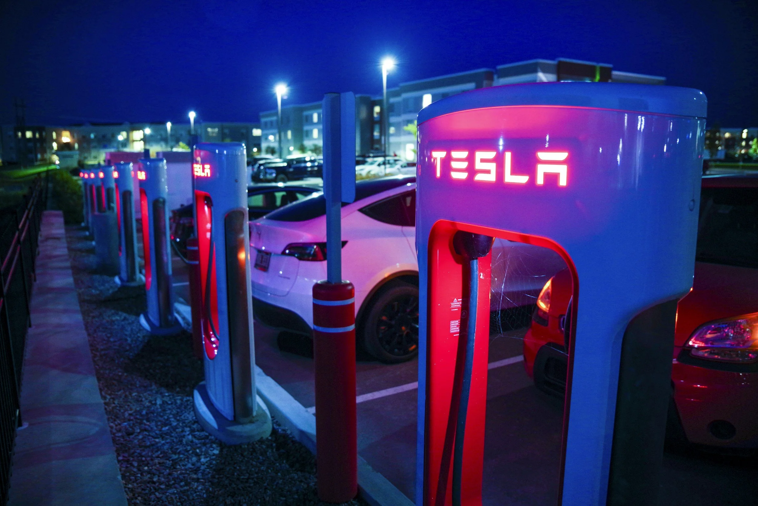 Tesla electric vehicle charging stations at night with cars parked nearby and residential buildings in the background.