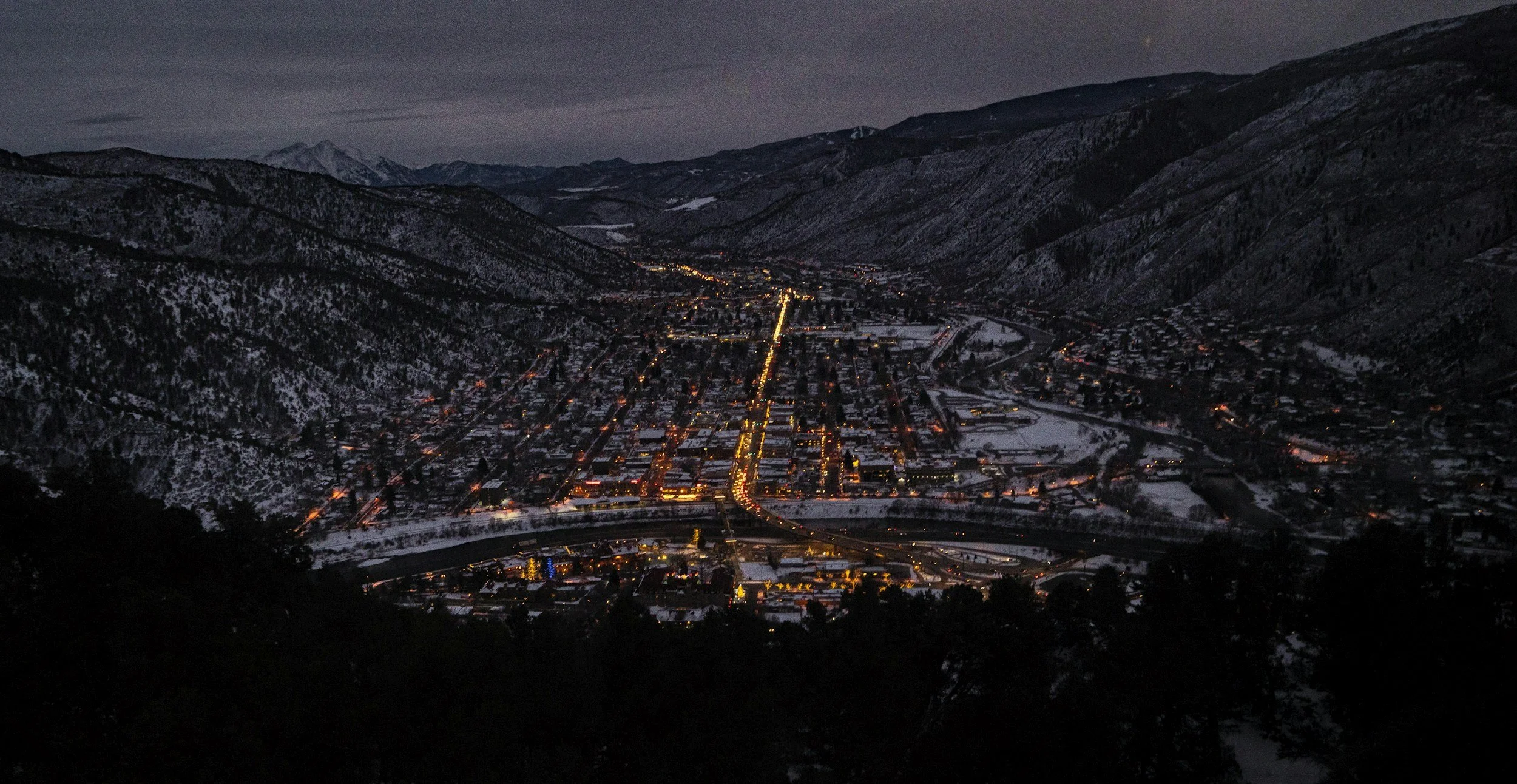 Nighttime view of a small town in a mountain valley with snow on the ground, illuminated by streetlights, surrounded by dark, forested mountains under a cloudy sky.