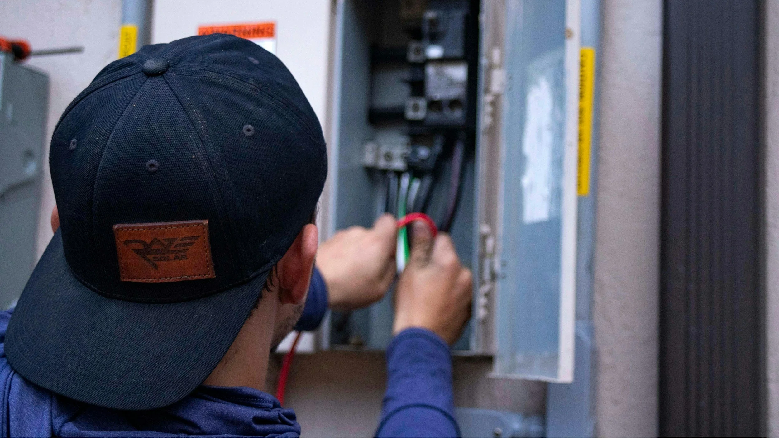 A person in a blue hoodie and black hat working on electrical wiring inside an electrical panel.
