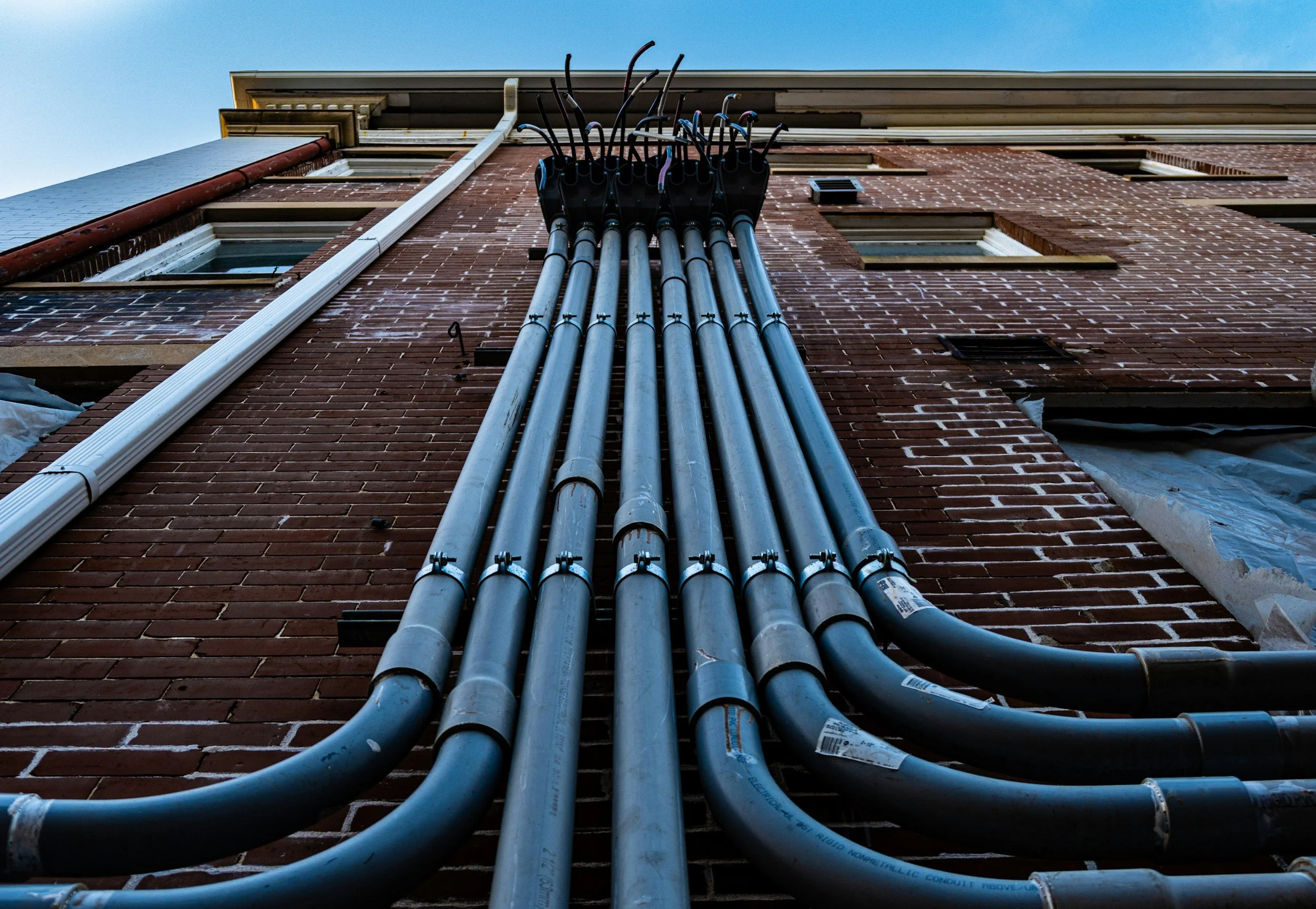 Multiple gray utility pipes running vertically along a brick building exterior with windows, under a clear blue sky.