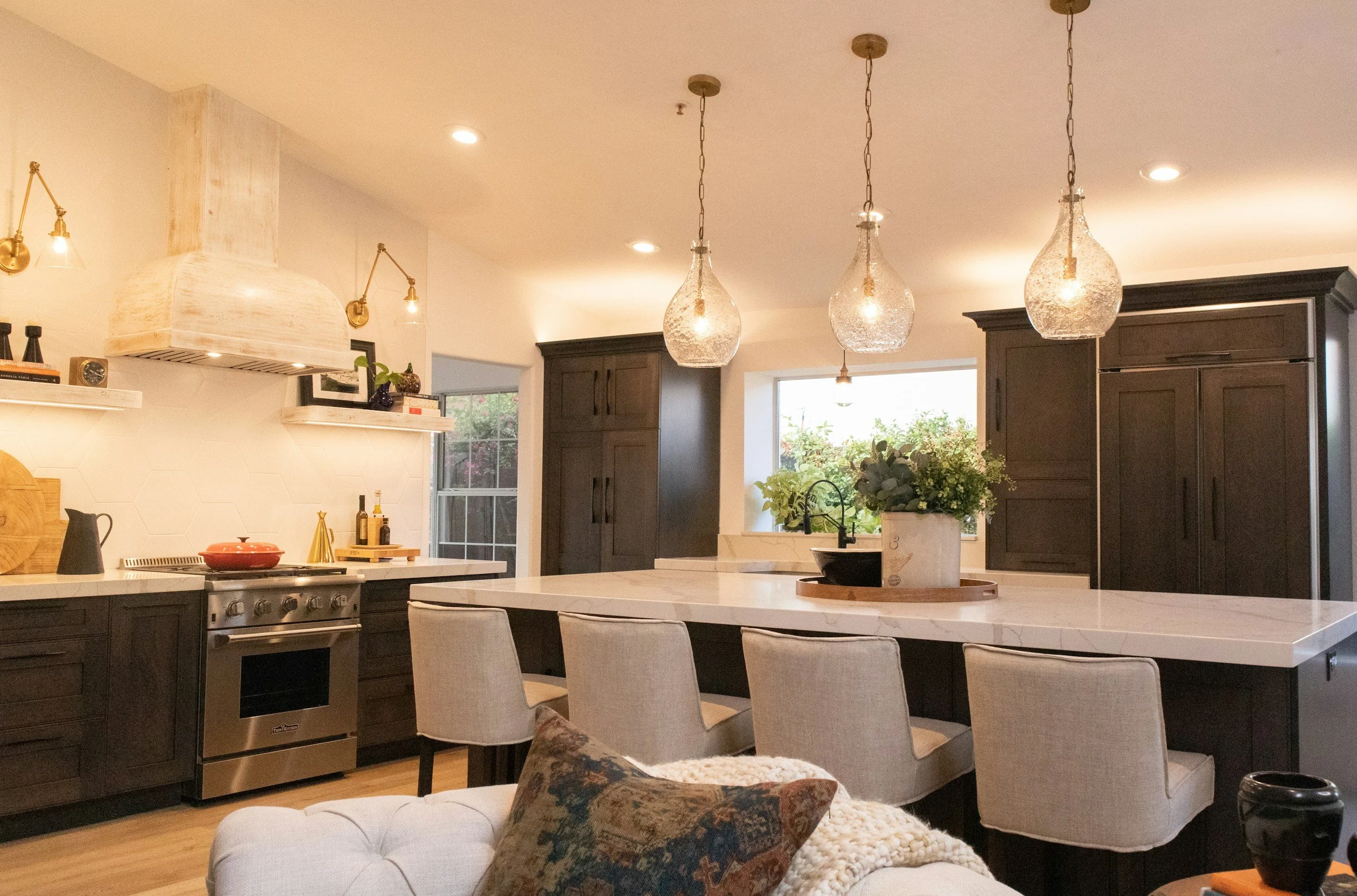 A modern kitchen with dark cabinets, a white island, pendant lights, and a cozy seating area with a cushion and throw blanket.
