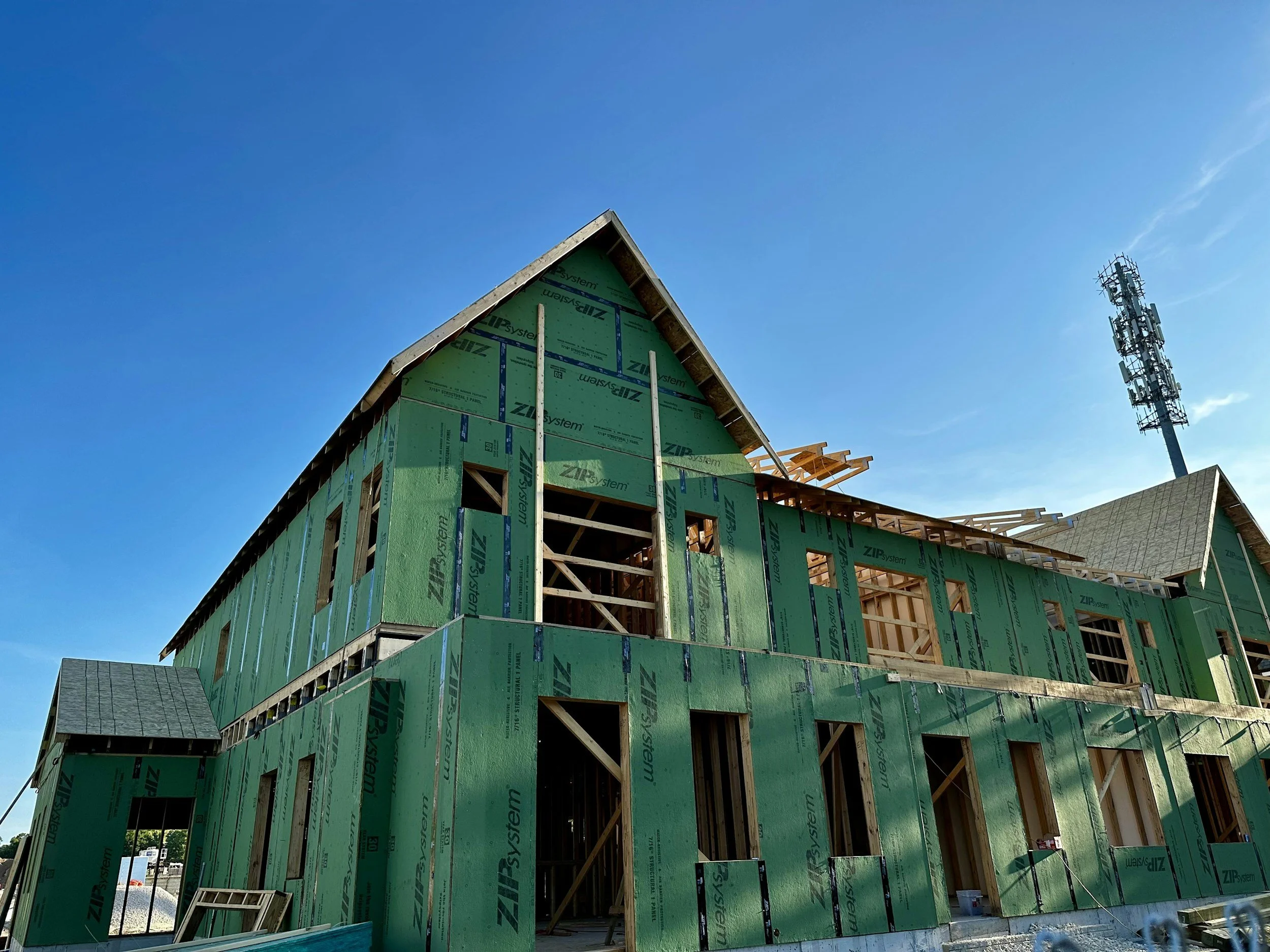 A building under construction with green insulation panels and exposed wooden framing, against a clear blue sky.