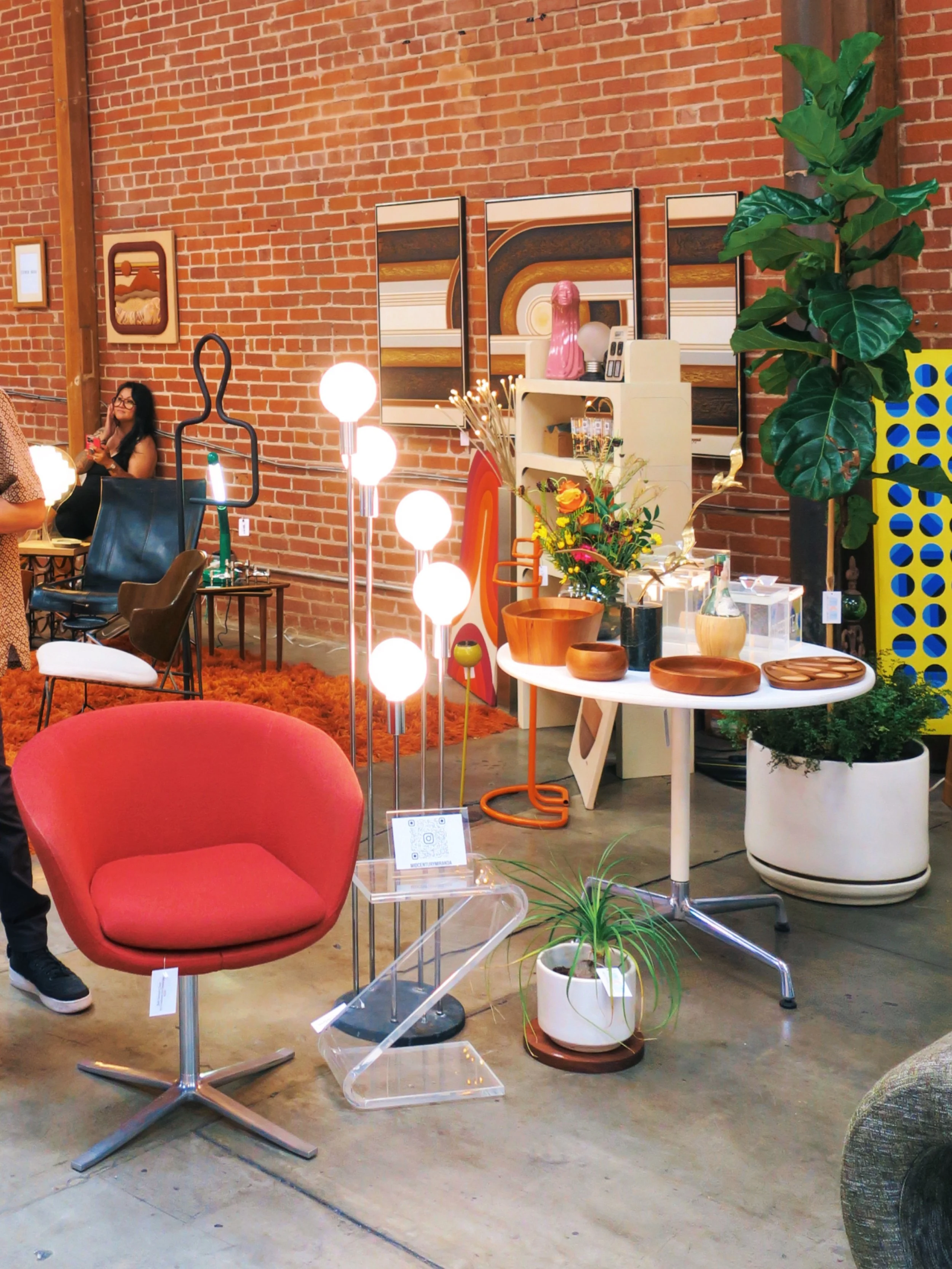 Display of mid century modern furniture and home decor items including a red Bob Swivel armchair, various wooden teak bowls, plants, large 1970s wall art, and vintage Robert Sonneman lighting inside a los angeles warehouse with exposed brick walls.