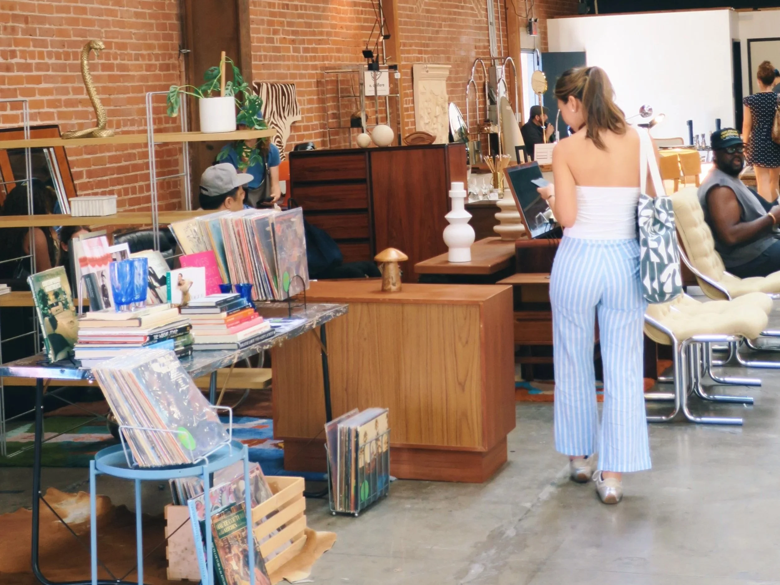 People shopping at a brightly lit vintage furniture market with brick walls, MCM furniture, books, vinyls, and decorative items, including a gold snake-shaped sculpture and a potted plant.