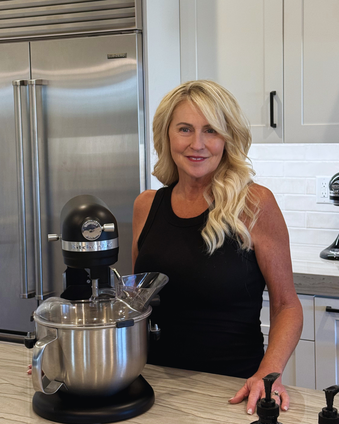 A woman with blonde hair in a black sleeveless top standing in a modern kitchen, smiling at the camera, with a stand mixer on the counter.