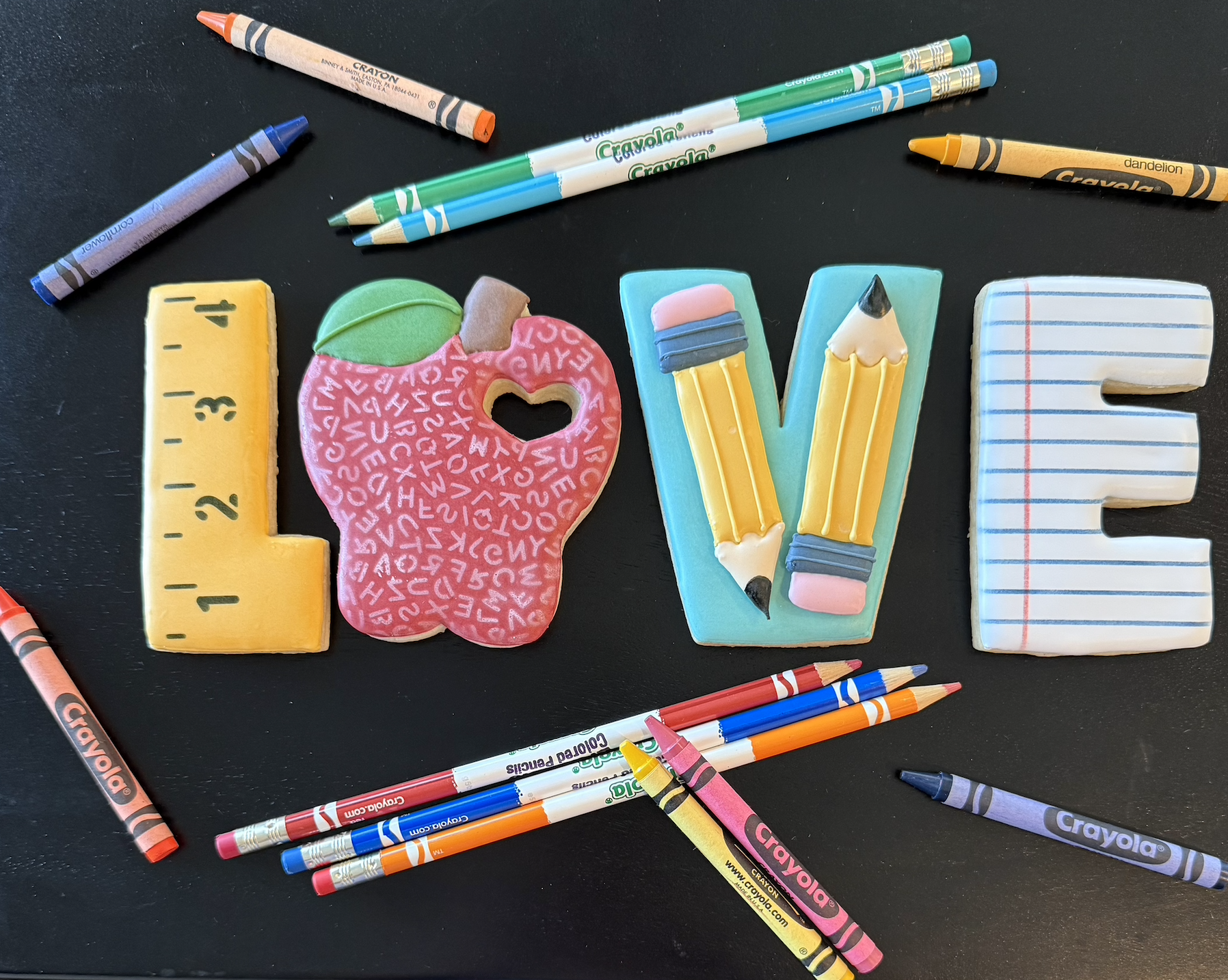 Colorful cookies shaped like words spelling out 'LOVE' with various classroom supplies around them, including pens and markers on a black surface.