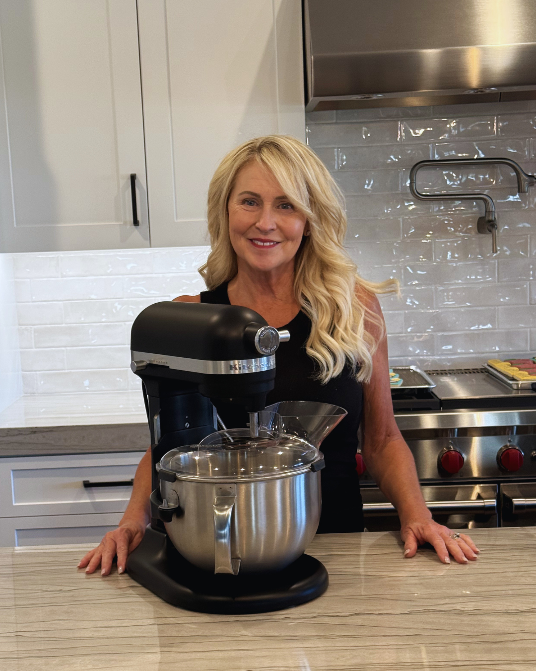 A woman with blonde wavy hair standing in a modern kitchen, smiling, with a stand mixer on the countertop in front of her.