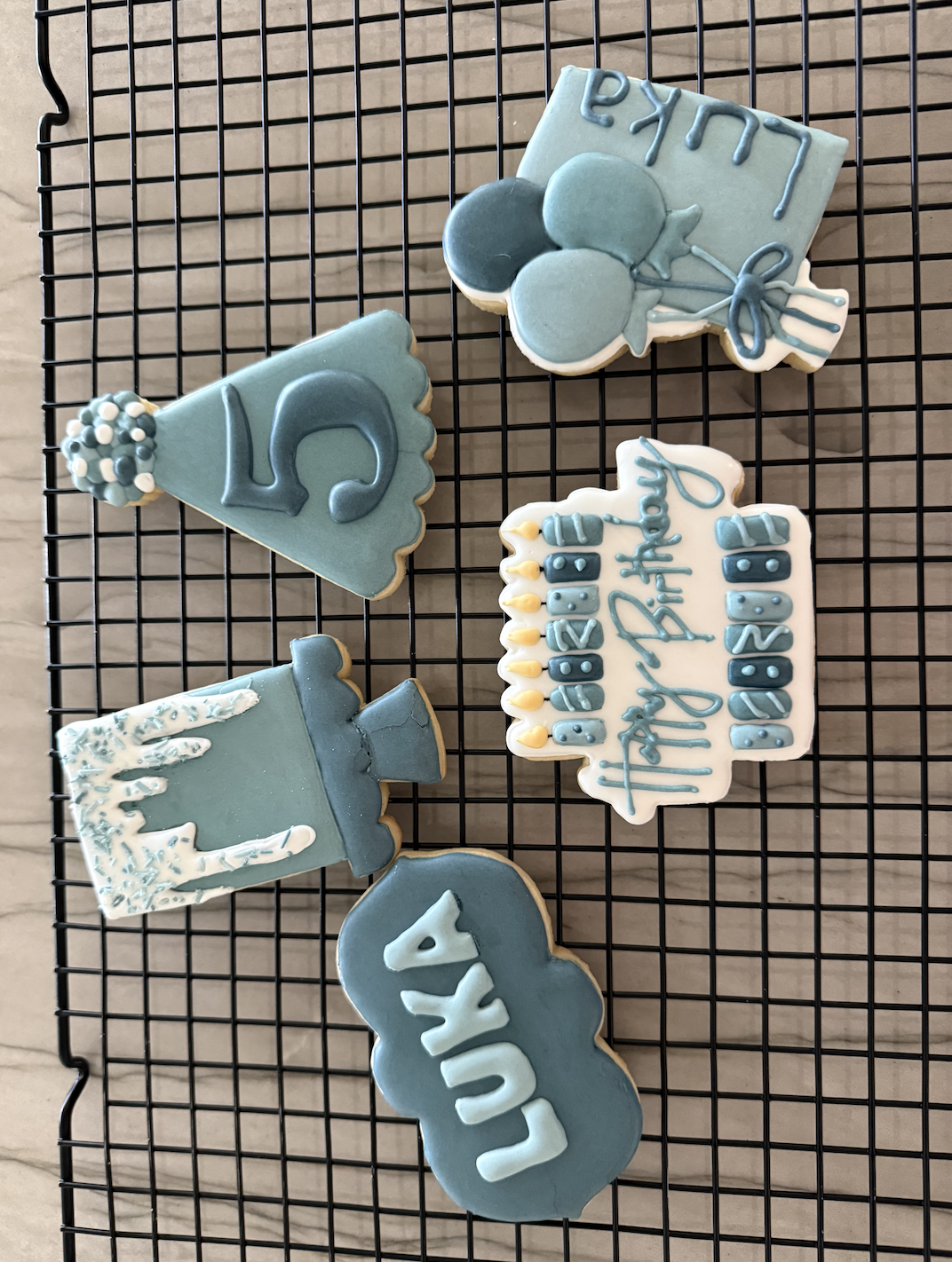 Set of six light blue and white birthday cookies on a cooling rack, decorated with balloons, a hat, a candle, and birthday messages.
