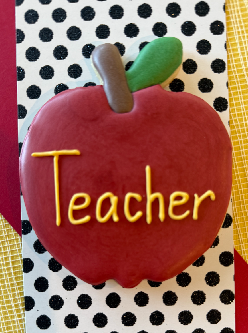 A decorative apple-shaped item with the word 'Teacher' written on it in yellow, green, brown, and red colors, placed on a polka dot surface with yellow and red accents.