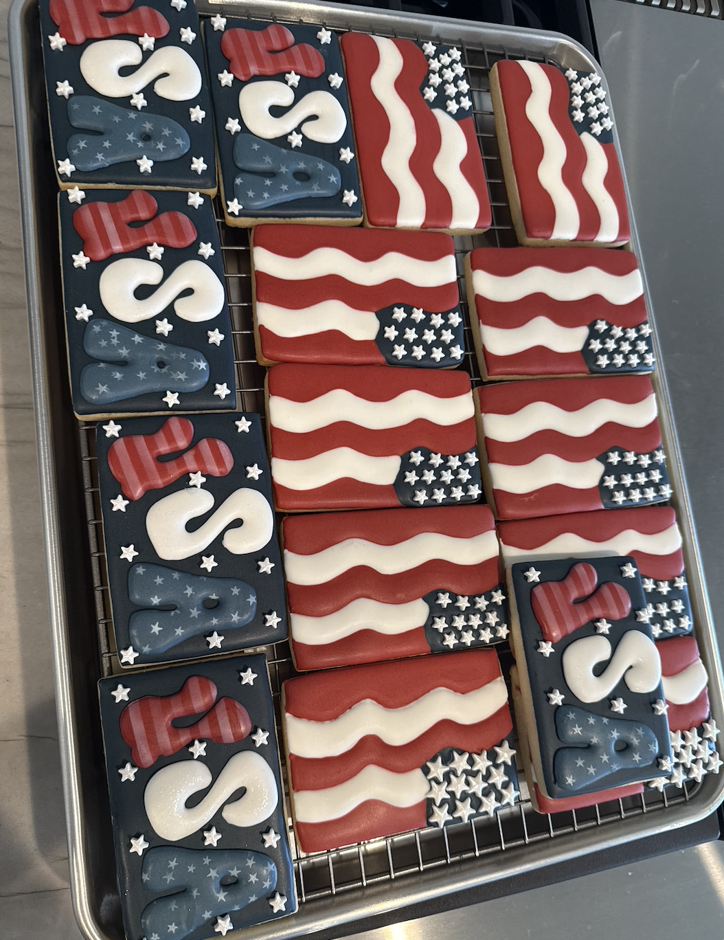Cookies decorated with American flag-themed patriotic designs, including red, white, and blue colors, stars, and stripes, arranged on a cooling rack.