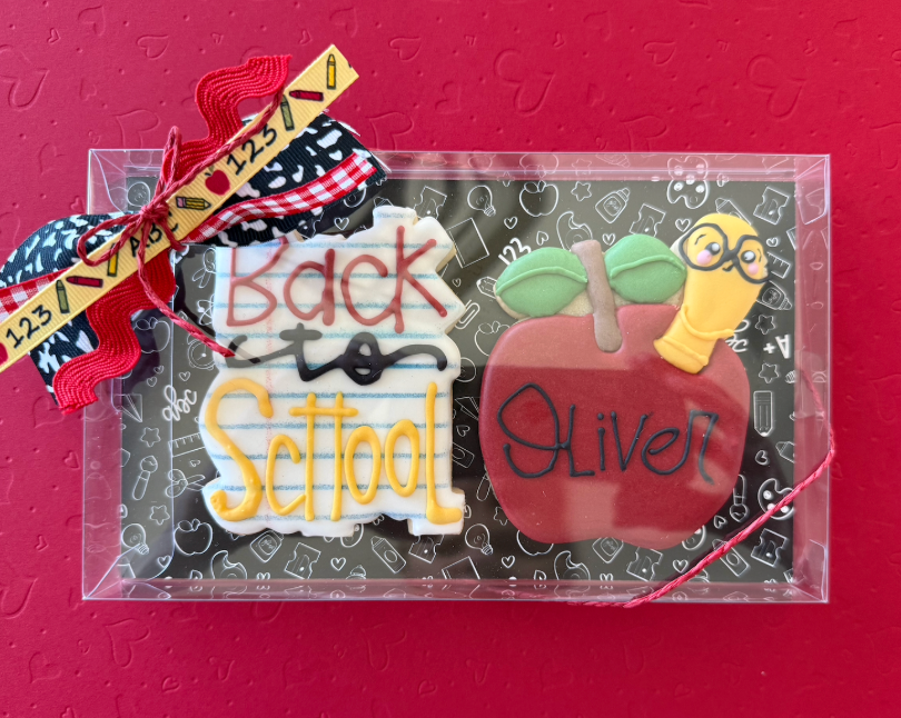 Decorated cookies in a clear box with a red background. One cookie reads 'Back to School' and the other is an apple with the name 'Oliver' and a worm with glasses.