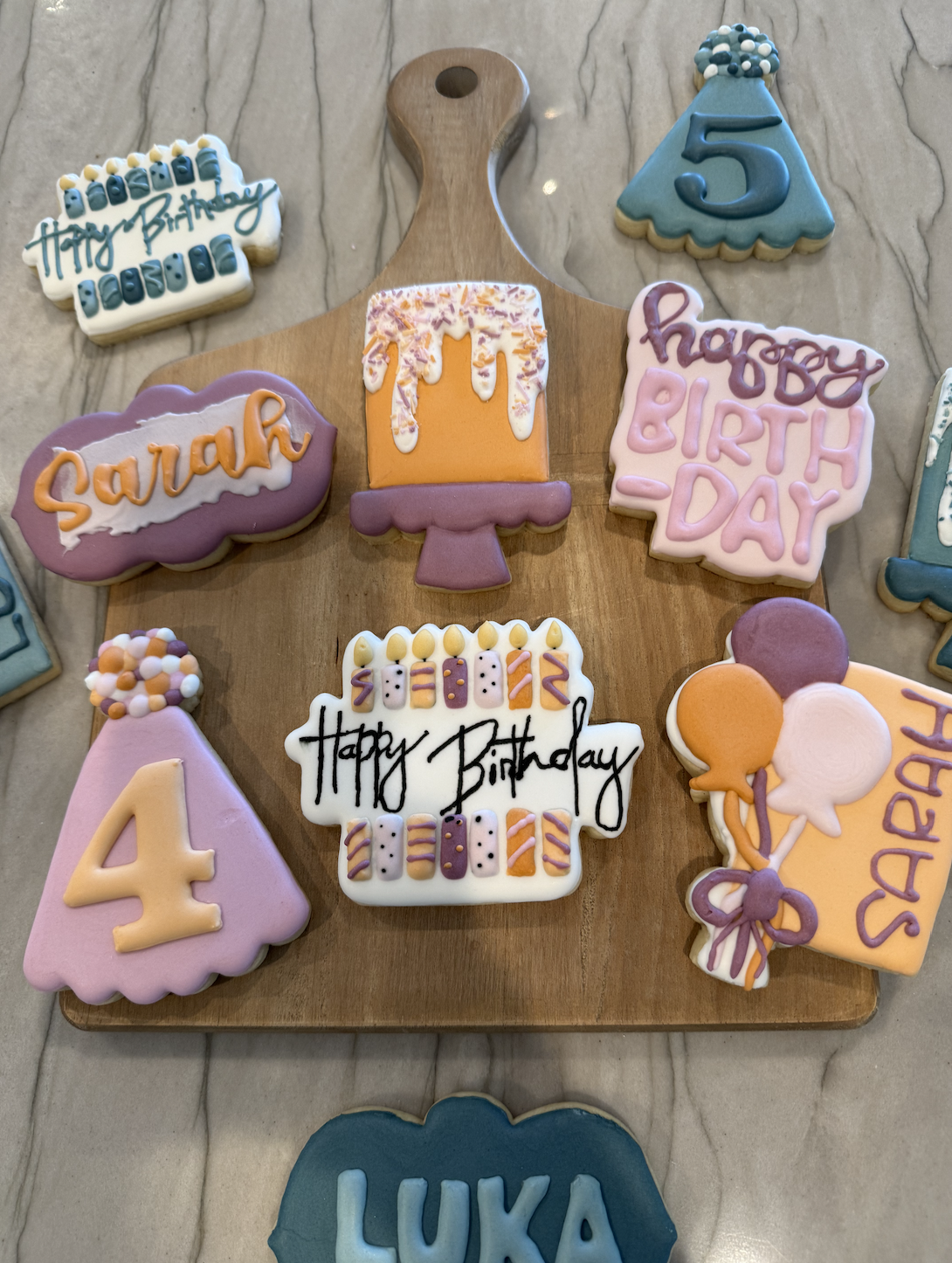 Colorful birthday cookies with various designs, including birthday messages, age numbers, and a party hat, arranged on a wooden cutting board on a marble surface.