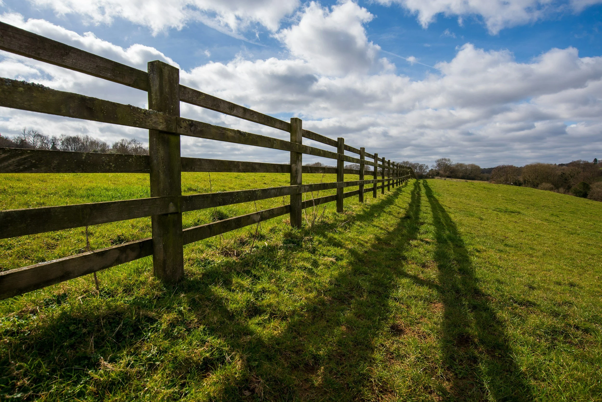 A weathered wooden fence lining a green grassy field under a partly cloudy blue sky.