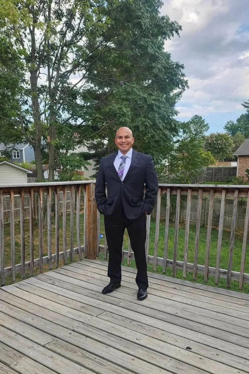 Man in a dark suit and striped tie standing on a wooden deck in a backyard with green grass, trees, and houses visible in the background.