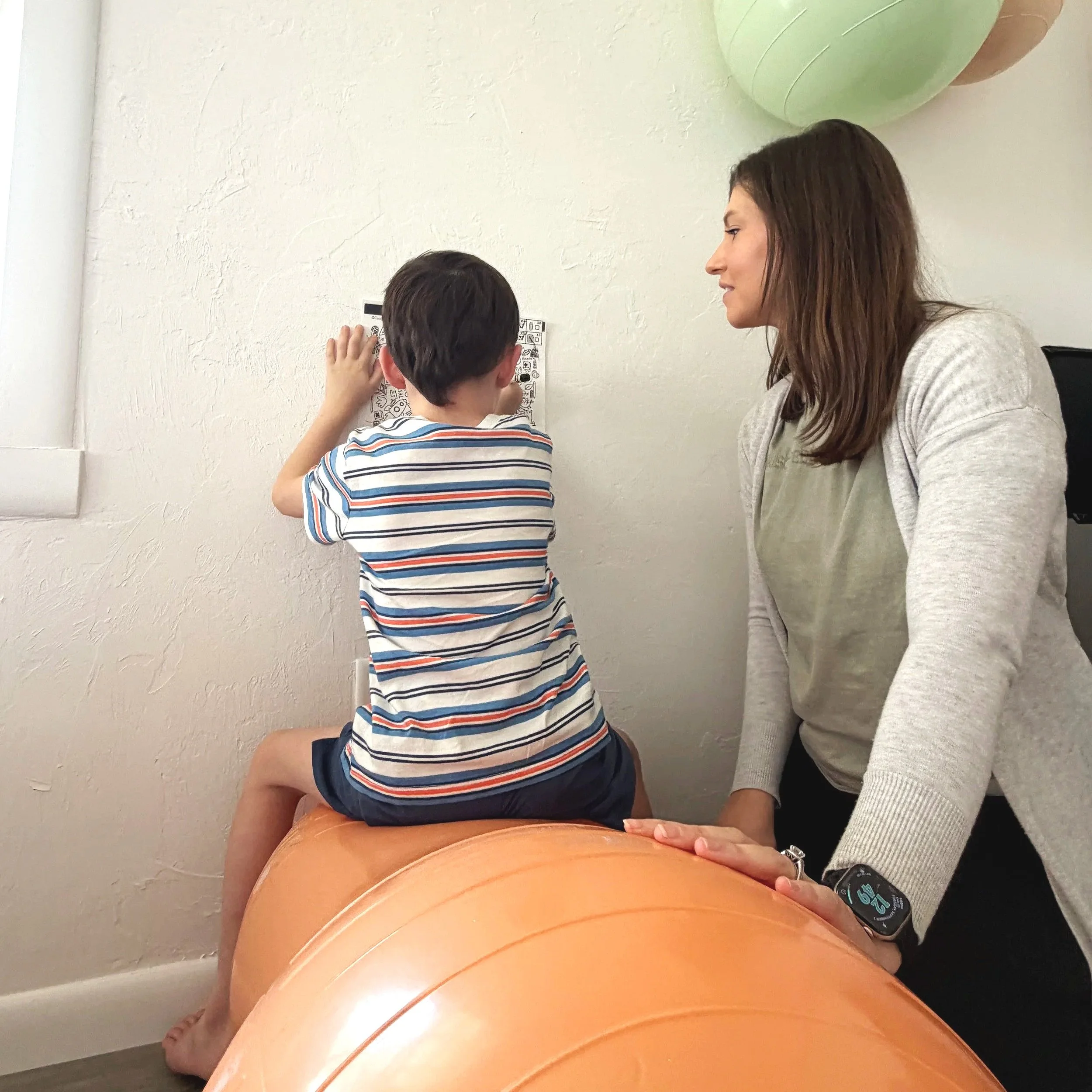 A pediatric occupational therapist and a young boy are sitting next to each other, with the boy touching a paper. They are seated at a table that has a large orange exercise ball nearby. She is addressing ocular motor  concerns