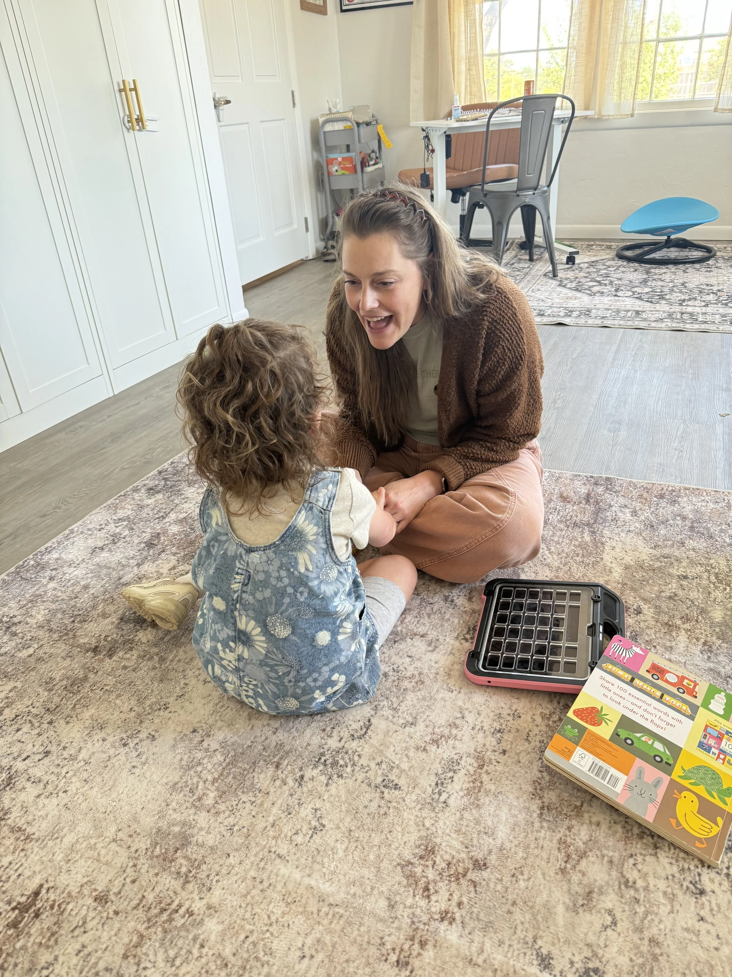 A pediatric speech therapist and a young girl sitting on a rug, facing each other, holding hands and smiling. Engaged in an AAC session addressing receptive and expressive language. There is a game box on the floor nearby.
