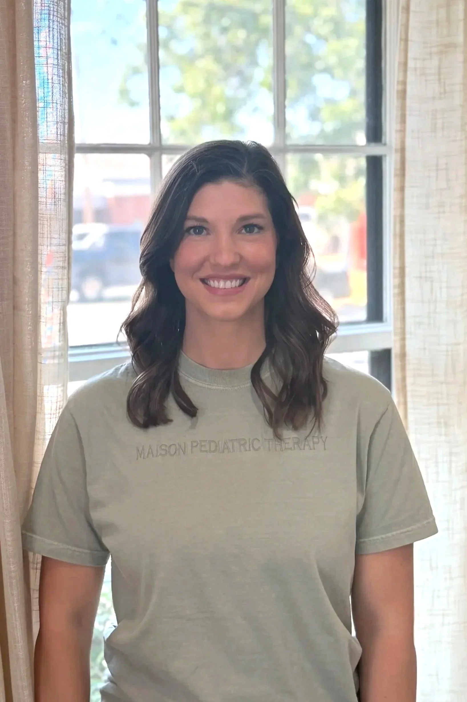 A woman with long dark hair smiling, wearing a light-colored t-shirt that says 'Maison Pediatric Therapy,' standing in front of a window with natural light.