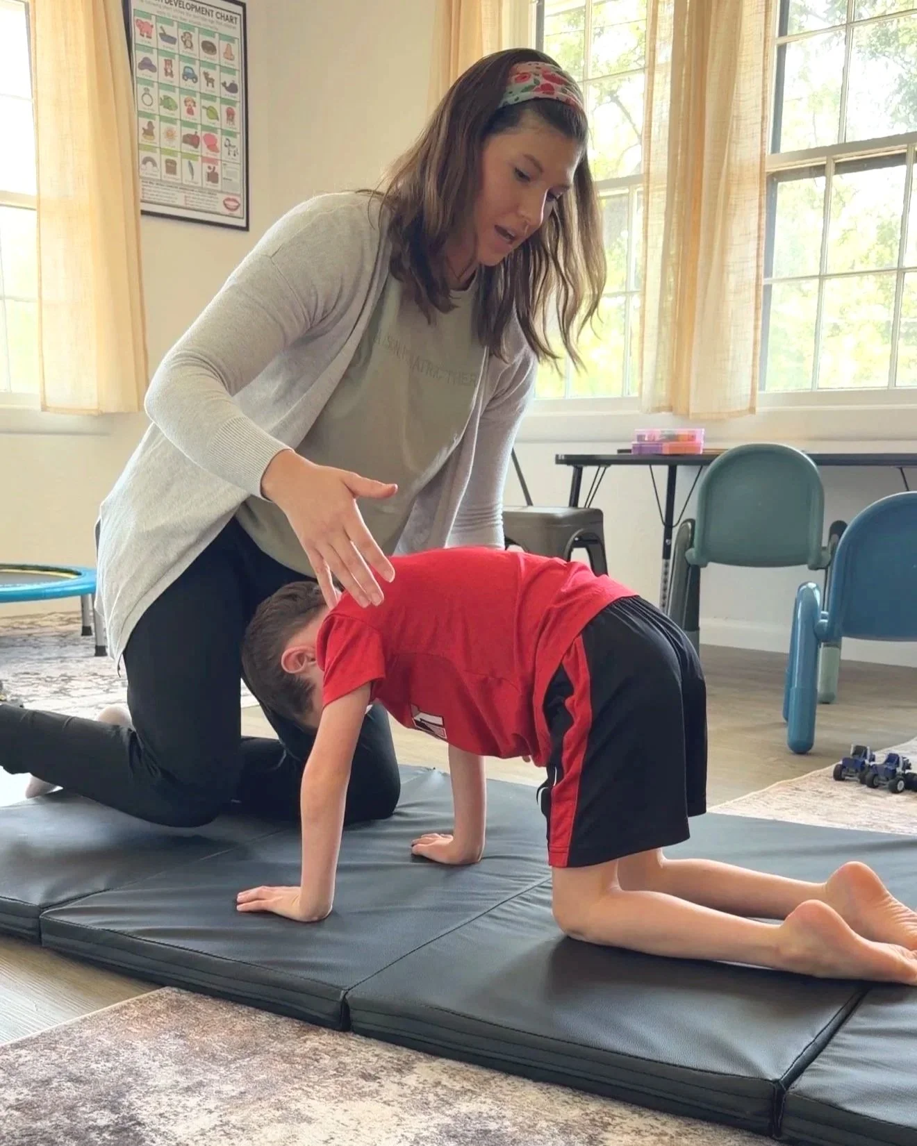 An occupational therapist in Edmond, Oklahoma evaluating a young boy in a Evaluation session, helping him perform a stretch on a black mat in a bright room with windows, chair and assessing for retained reflexes