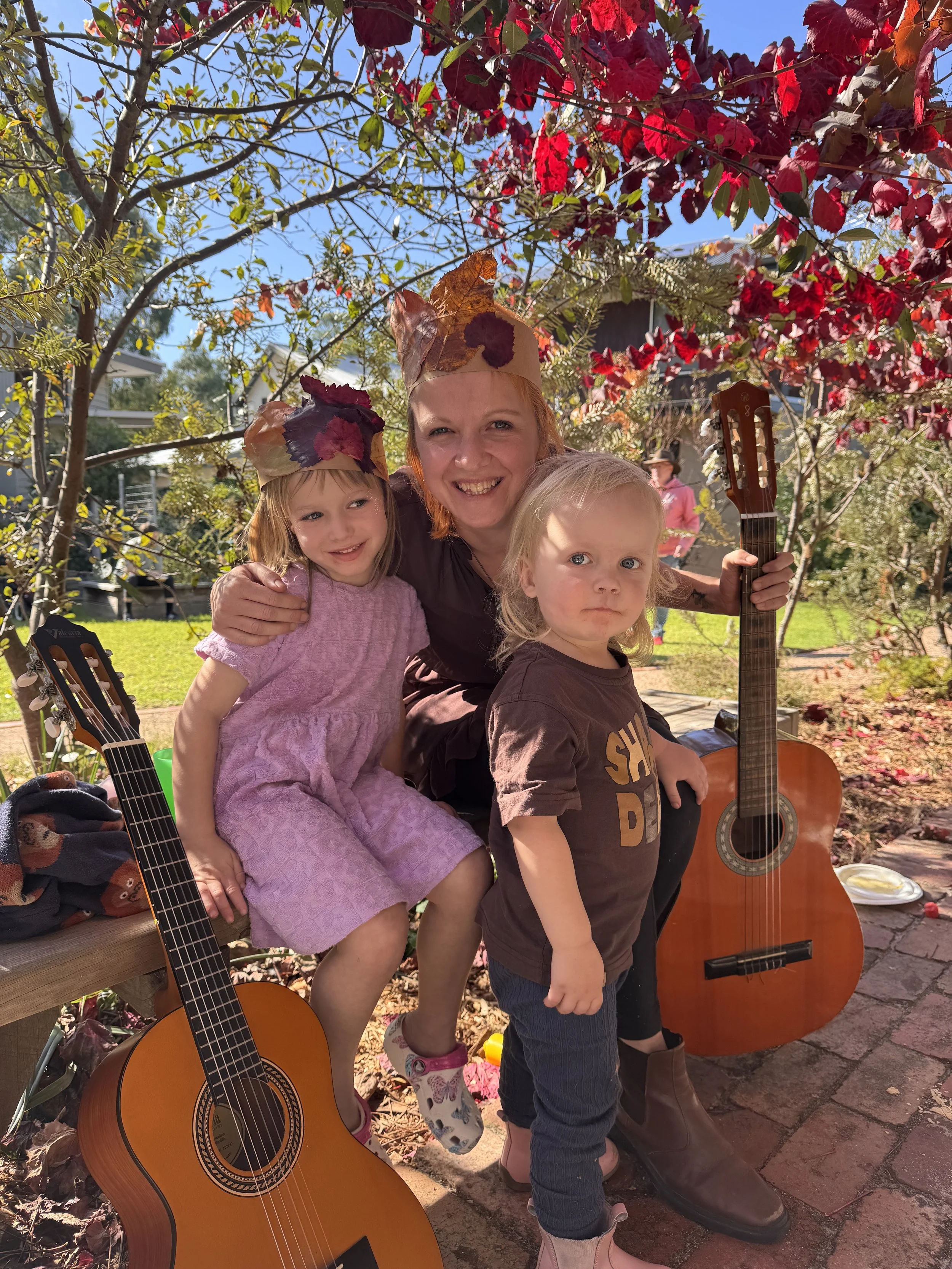 A woman with two young children outdoors during autumn, wearing leaf crowns, smiling, with guitars around them, surrounded by colourful fall foliage.