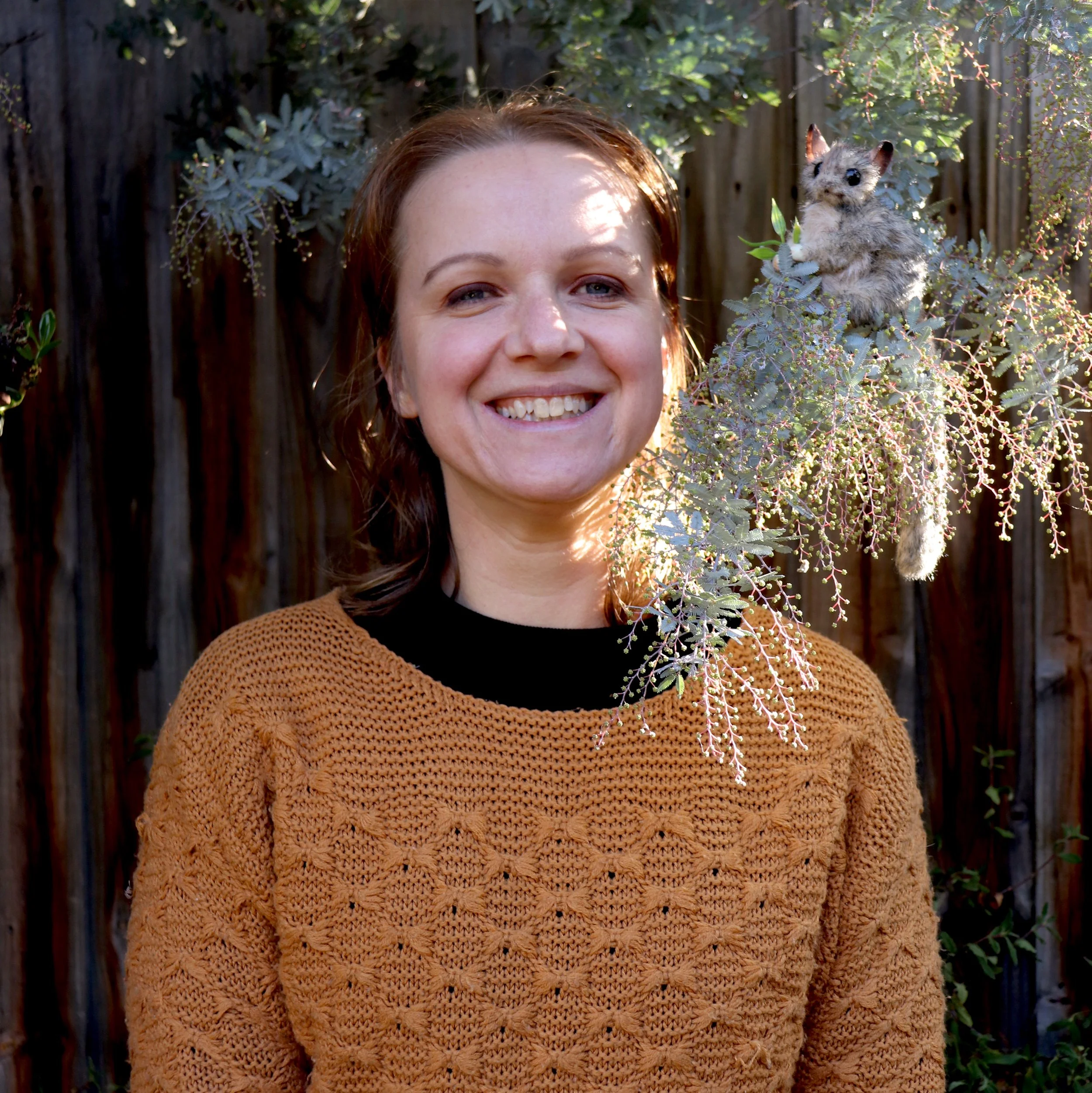 A woman with red hair smiling outdoors next to a bush with a small furry animal that has large ears, resembling a fairy possum, perched on the bush near her face.