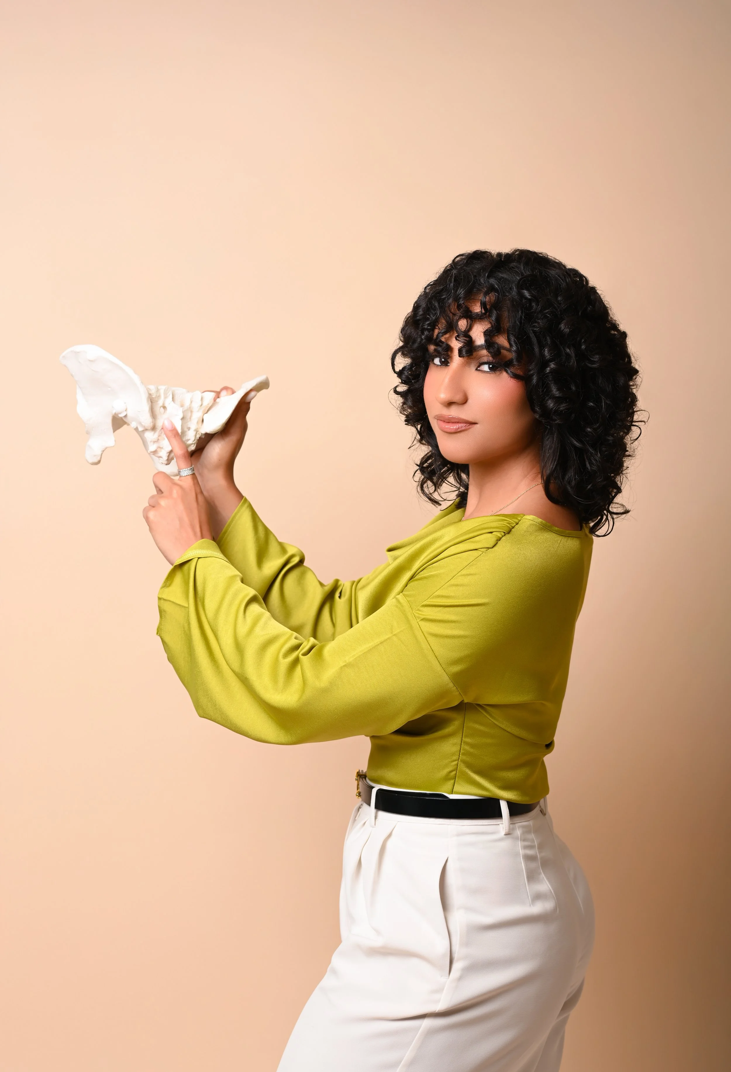 A woman with curly black hair holding a white sculpture of a bone, wearing a green blouse and white pants, standing against a beige background.