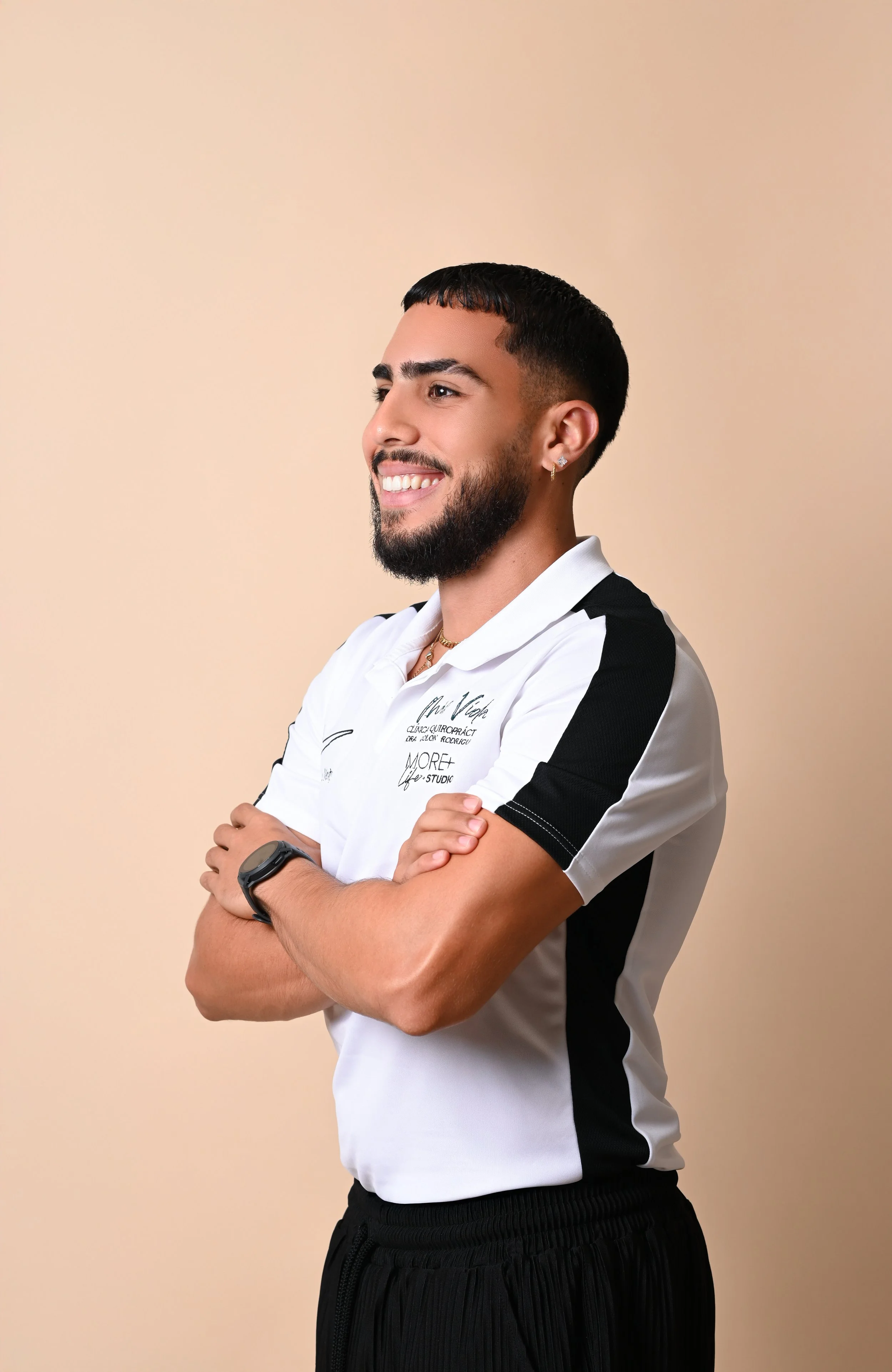 Smiling man with dark hair and beard, wearing a white athletic polo with black accents, black joggers, and a smartwatch, standing with arms crossed against a beige background.