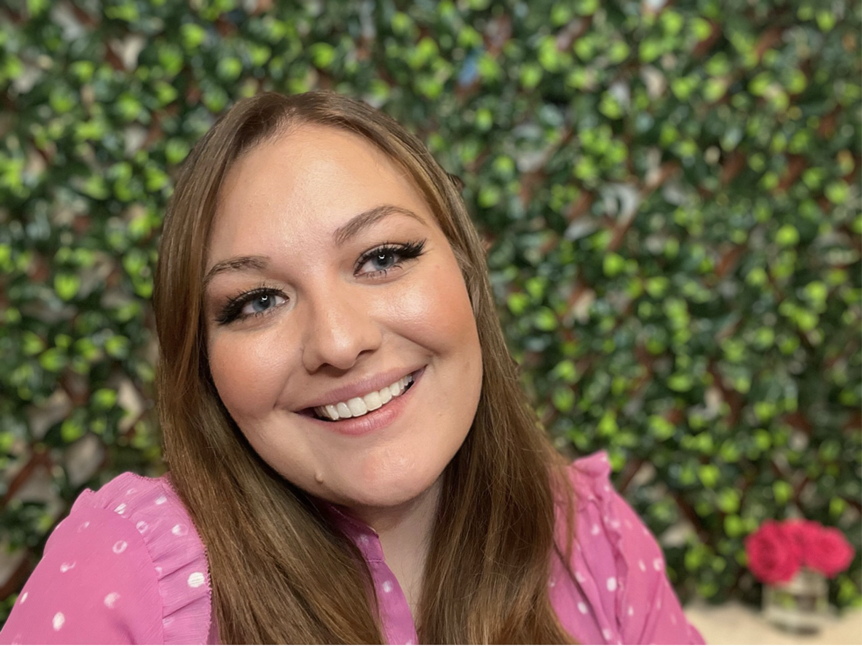 A smiling woman with long brown hair, blue eyes, and light skin, wearing a pink dress with white dots, standing in front of a background of green foliage and pink flowers.