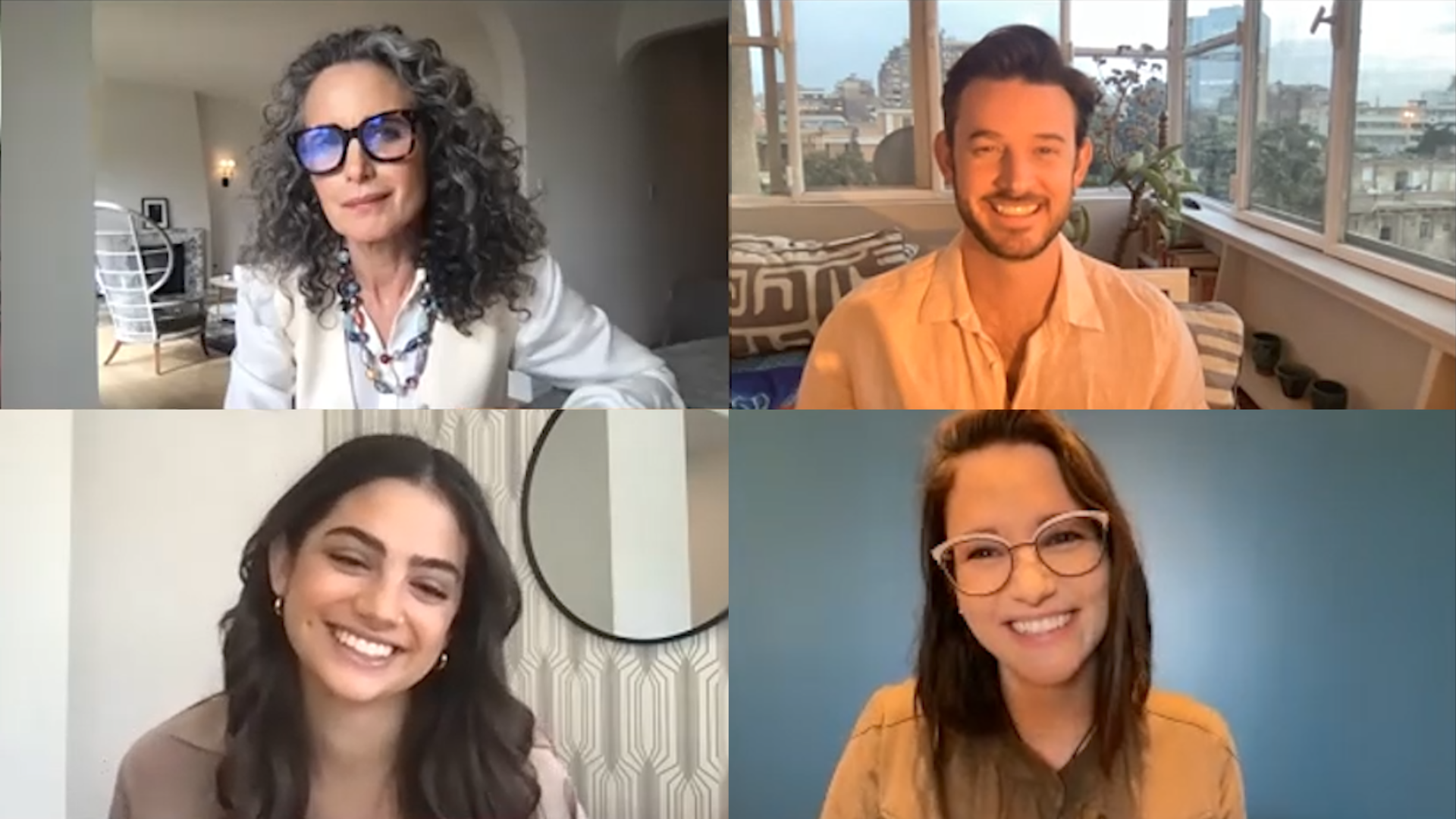 Four people in a virtual video call, two women at the bottom, one woman at the top left, and one man at the top right. The woman at the top left has curly gray hair, glasses, and is wearing a white blazer. The man at the top right has dark hair, a beard, and is wearing a peach-colored shirt. The woman at the bottom left has long dark hair and is smiling. The woman at the bottom right has glasses, long hair, and is wearing an orange top. They are all smiling and appear in well-lit indoor settings.