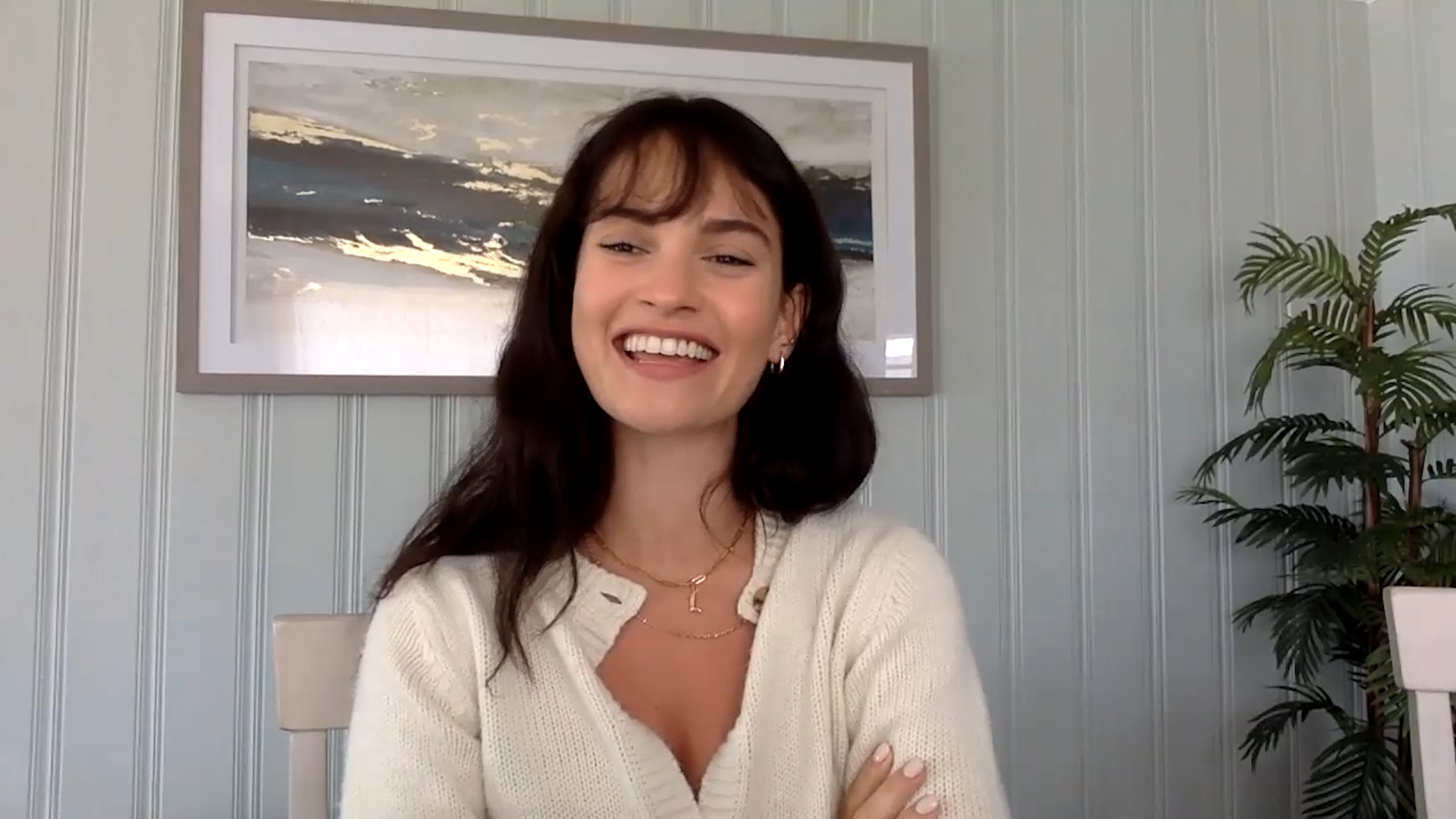 A woman with dark hair smiling and crossing her arms, seated in front of a light-colored paneled wall with a large framed painting of a sunset or sky scene and a green potted plant.