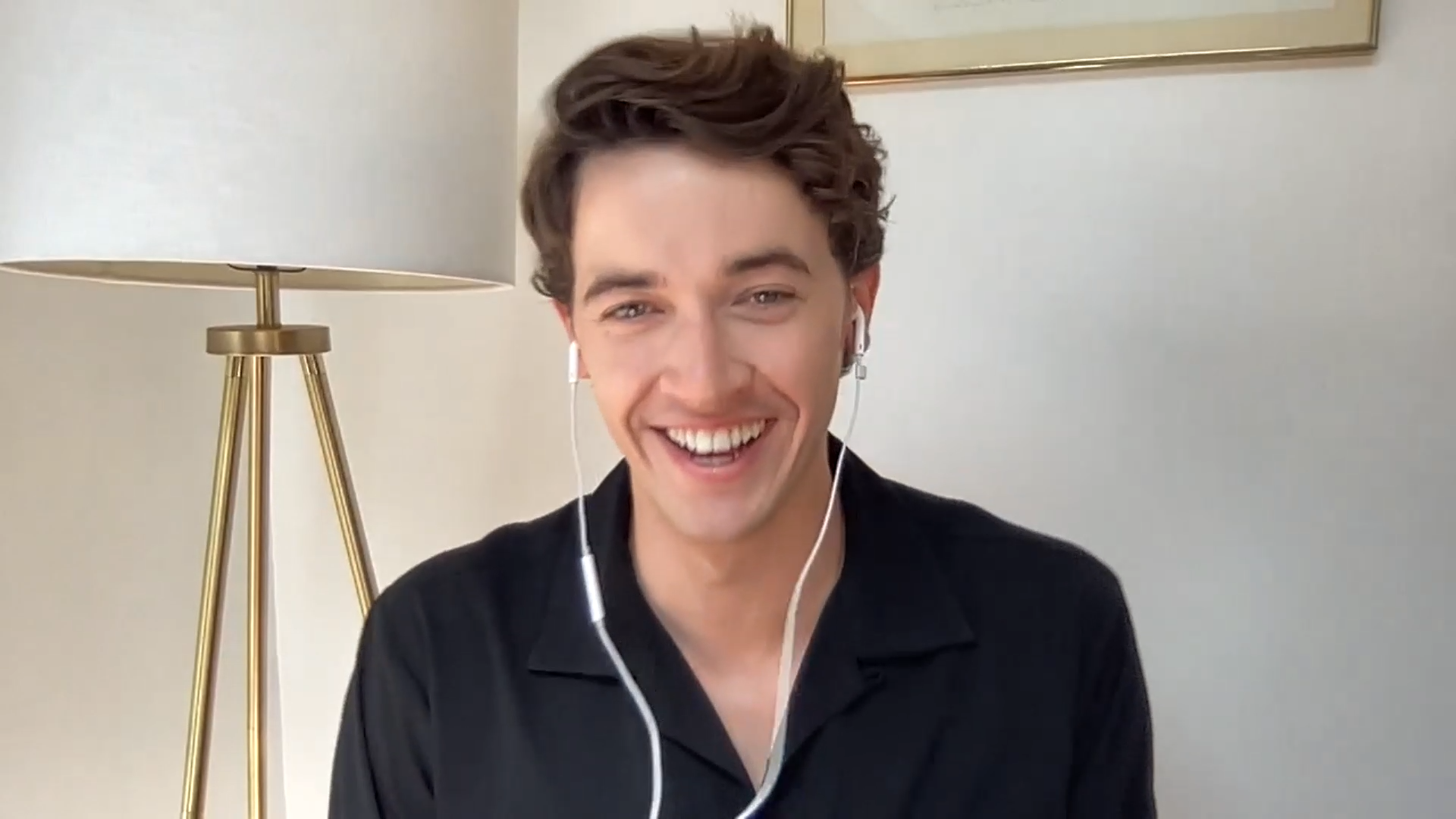 Smiling young man with brown hair wearing earphones, sitting in front of a white wall and a beige framed artwork, next to a white shade lamp with a metallic stand.